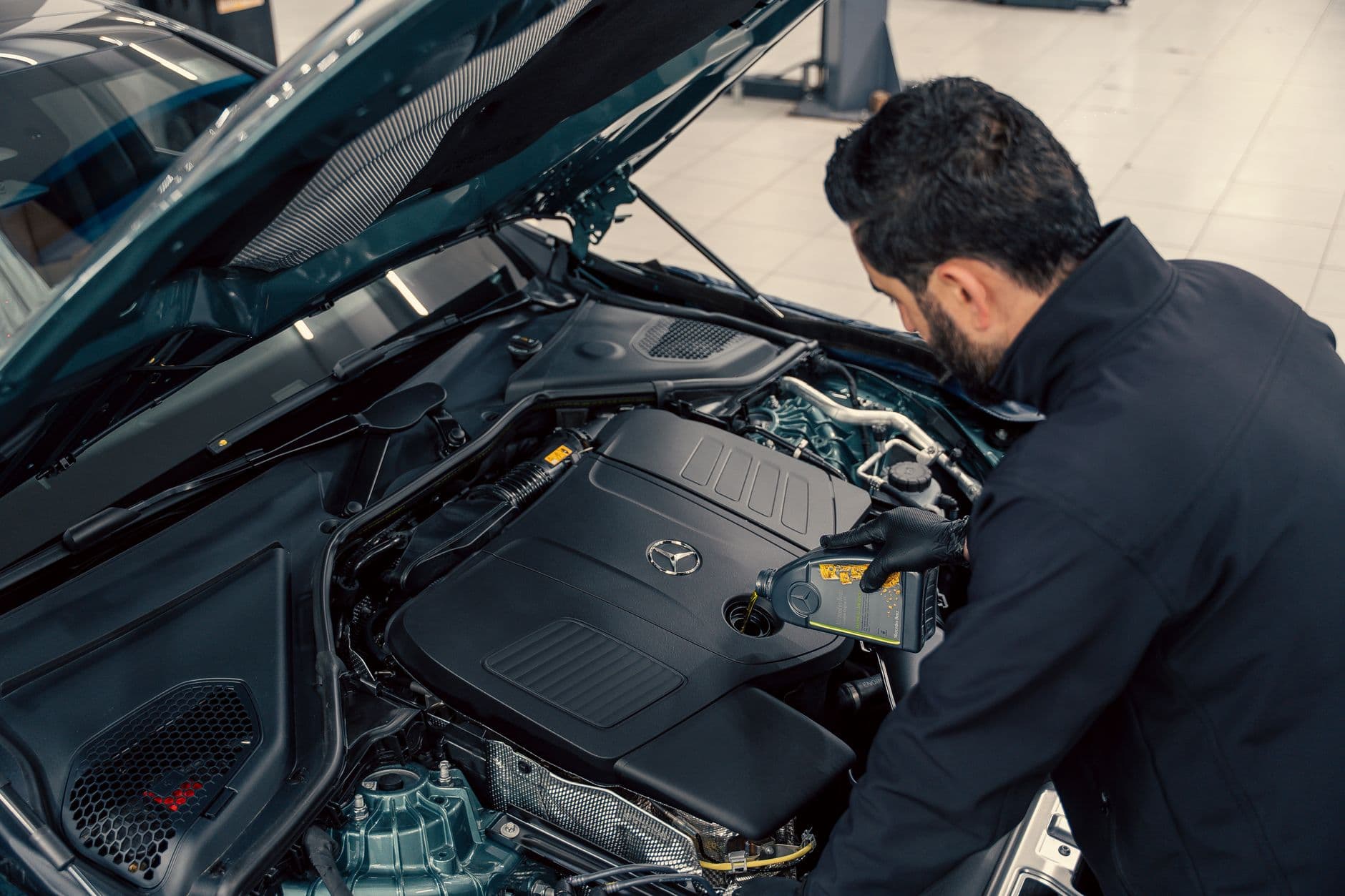 Mecanic care toarna ulei de motor dintr-un bidon in motorul unei masini cu capota ridicata, intr-un service auto