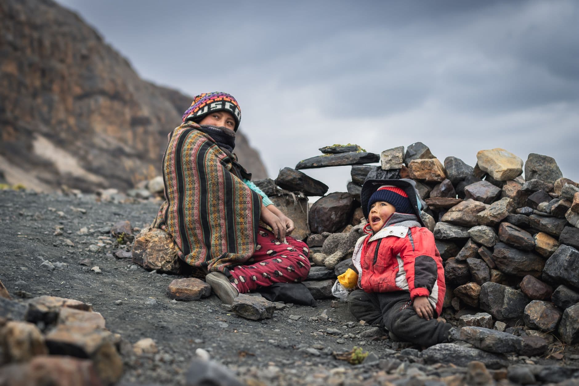 Mama si copil imbracati gros, asezati pe teren stancos in muntii Anzi din Peru, sub un cer noros