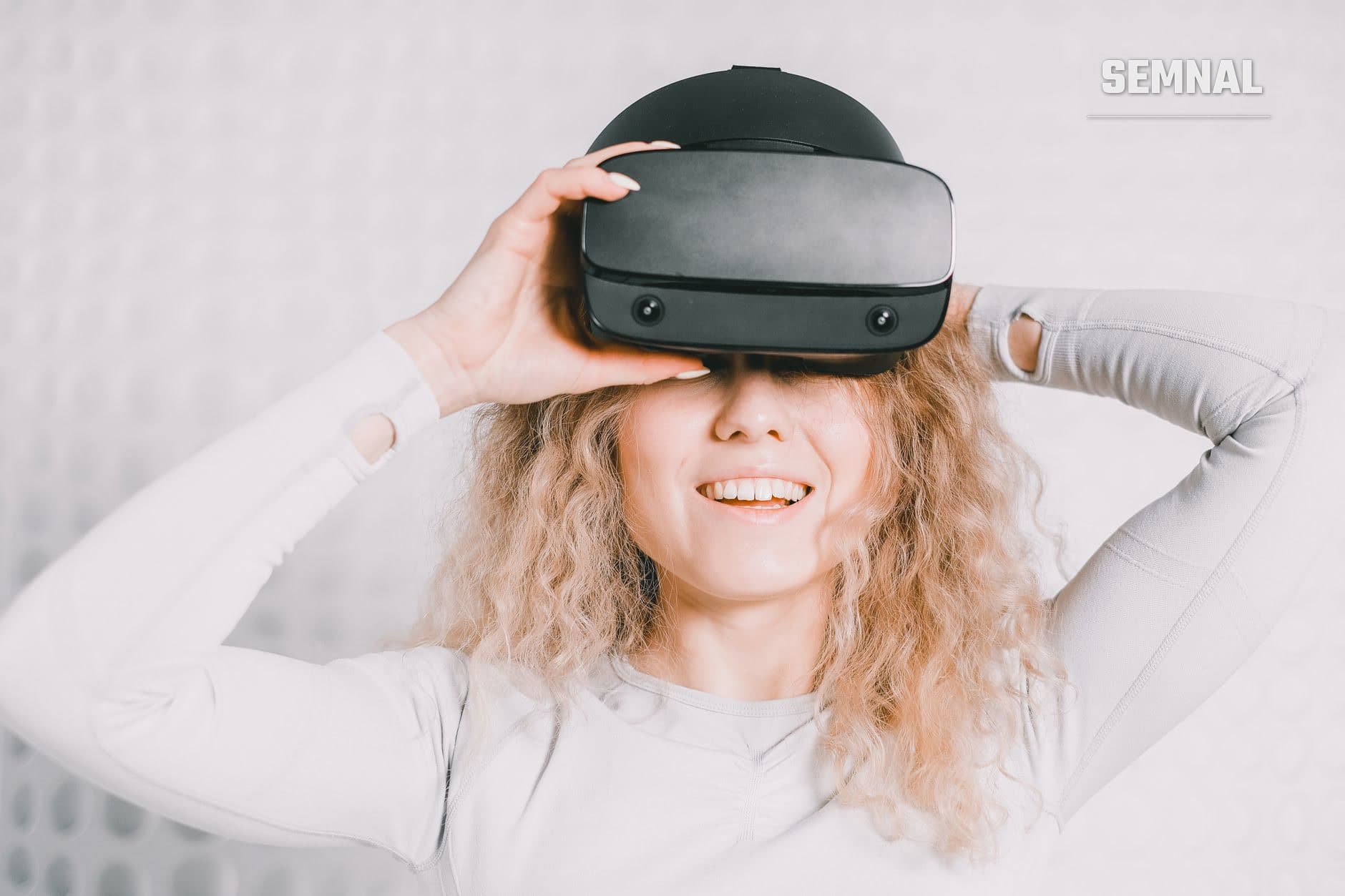 Persoană zâmbind ținând o cască de realitate virtuală neagră lângă capul cu părul cărunt, în cămașă albă, pe fundal alb.