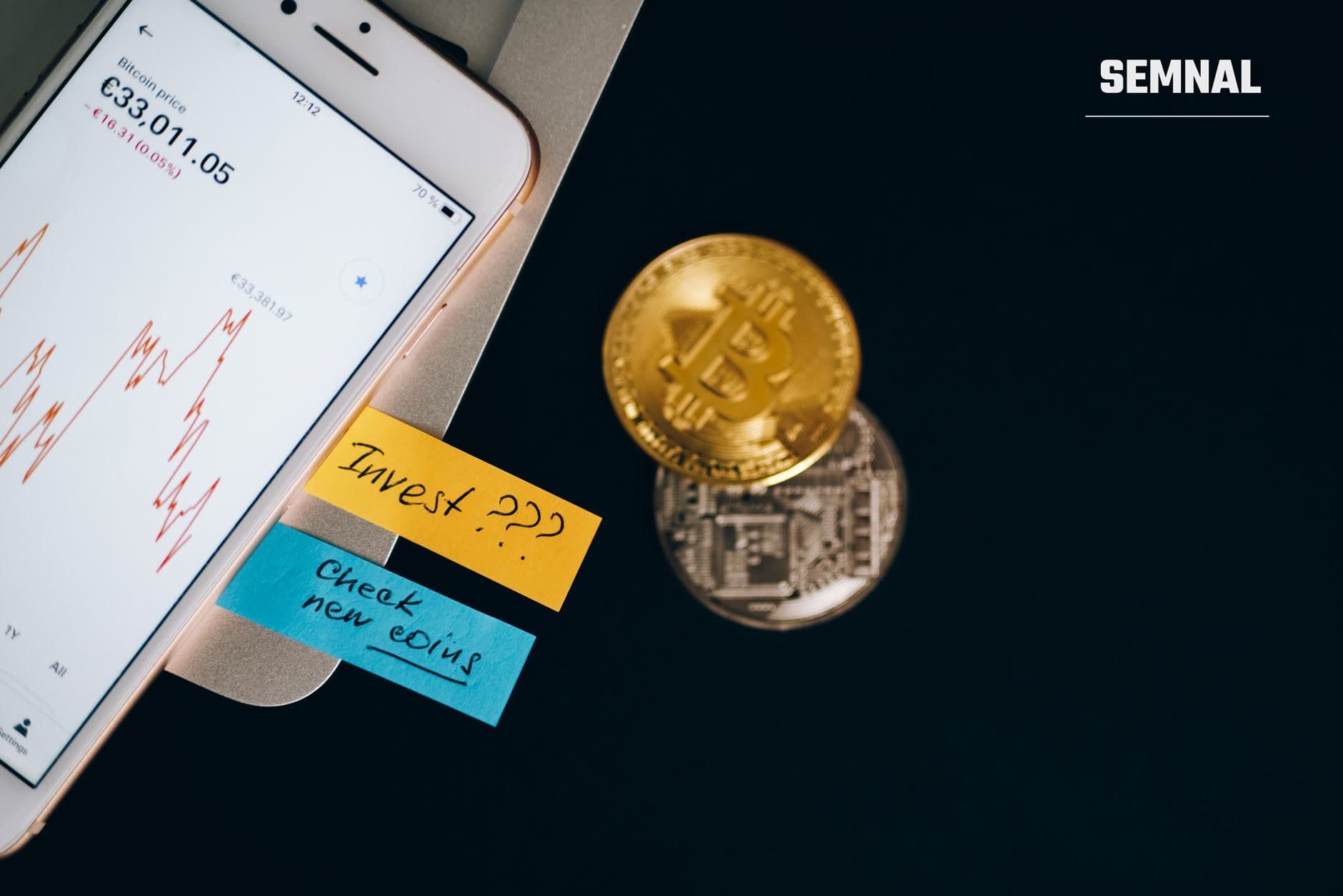 Telefon mobil cu grafic de preț și notițe adezive galbene și albastre, alături de o monedă bitcoin aurie pe fundal negru, ilustrând analiști și investiții în criptomonede.