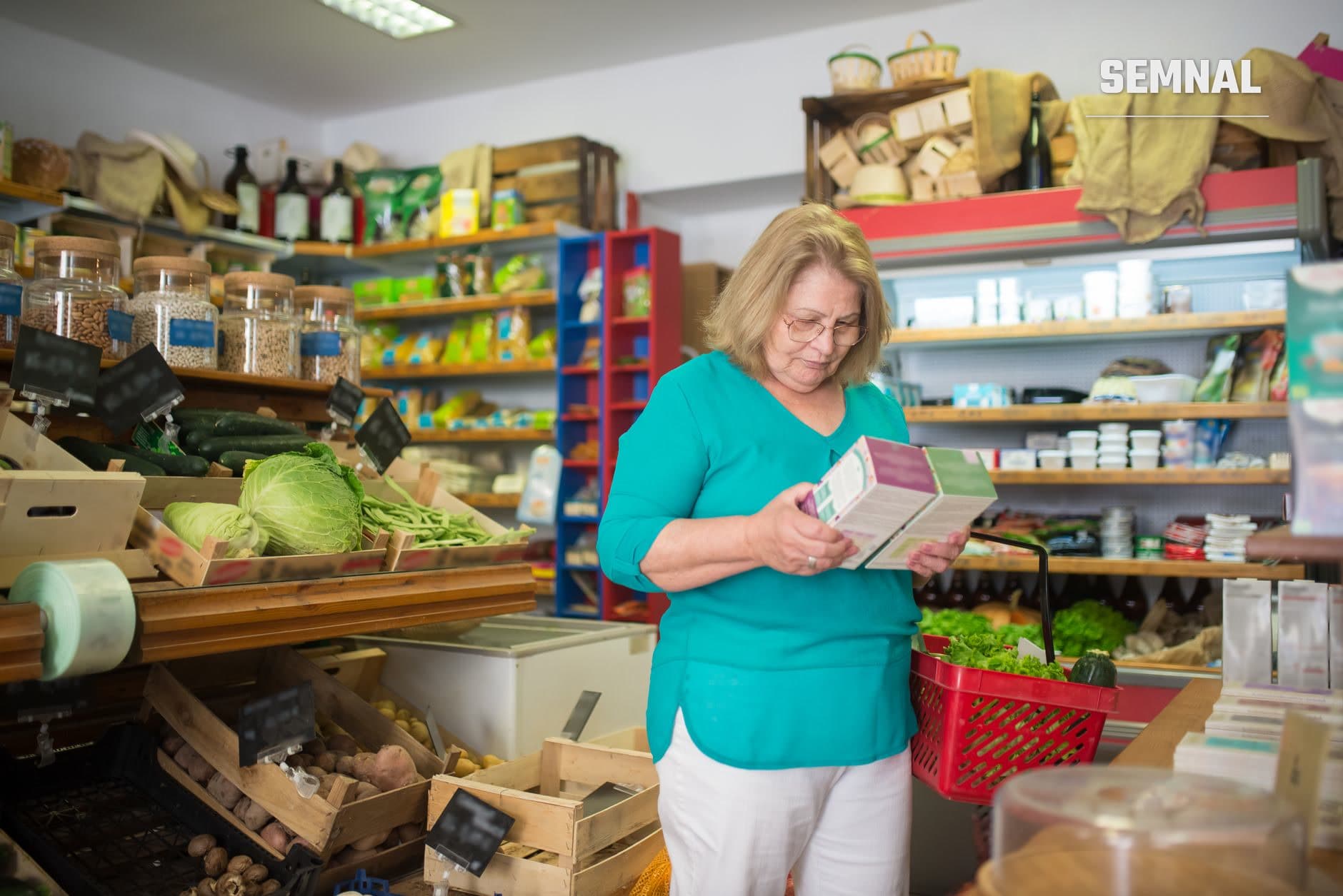 O femeie cu ochelari citește etichetele nutriționale ale unui produs alimentar într-un magazin de alimente, cu rafturi pline de produse și legume proaspete în fundal.
