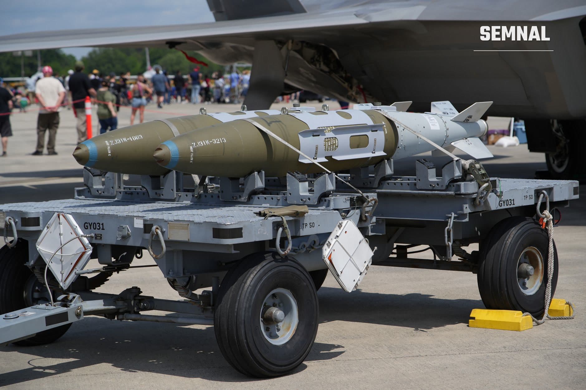 Două rachete aer-aer montate pe platformă de transport militar, una de culoare kaki și cealaltă gri-albă, expuse într-o demonstrație militară cu avion de transport în fundal.