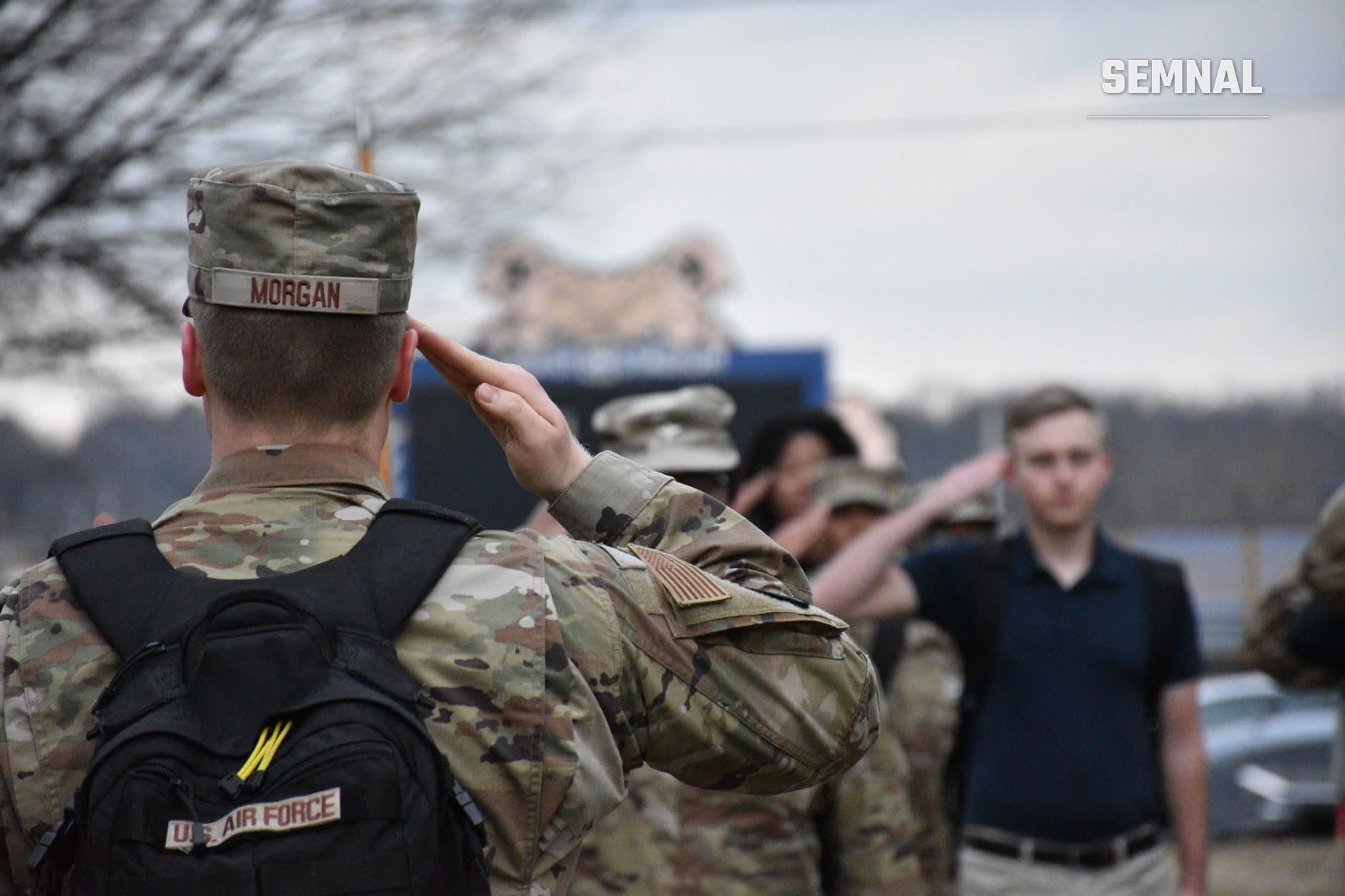 Militar american în uniformă de camuflaj cu insigna US Air Force salutând, purtând pălărie cu numele 'Morgan', cu alți soldați și civili în fundal la o ceremonie militară.