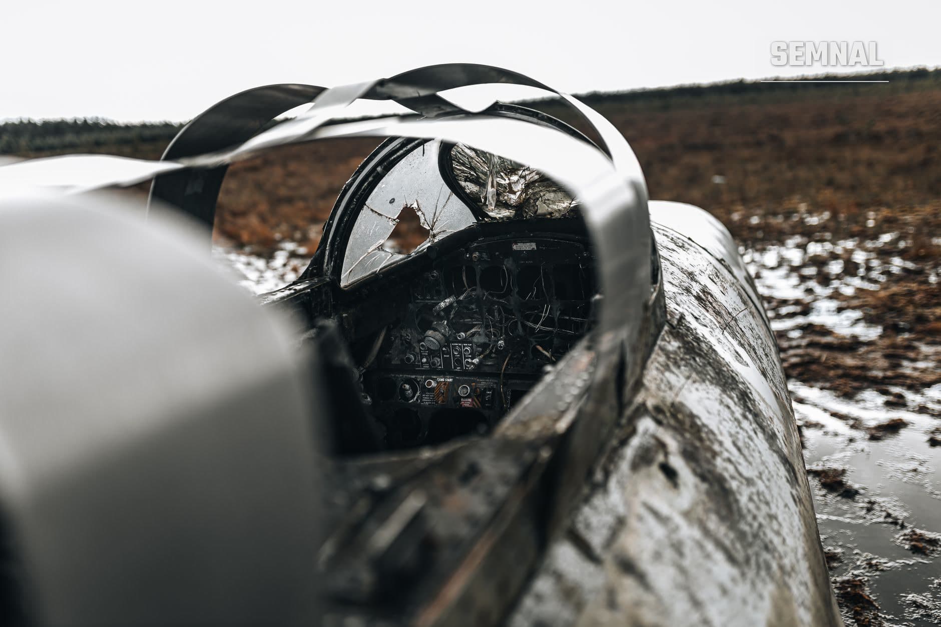 Epavă de avion militar cu fuselaj alb și negru, oglindă ruptă și cockpit distrus pe teren neigienic cu zăpadă.