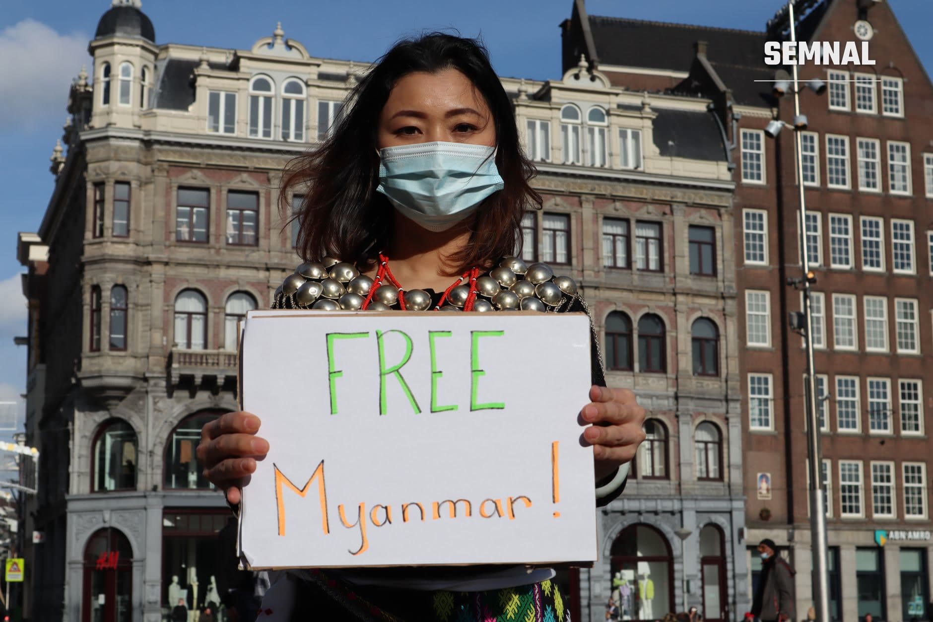 O tânără în mască protectoare ține o pancardă cu mesajul 'FREE Myanmar!' în fața unor clădiri istorice, participând la un protest pentru drepturile omului în Myanmar