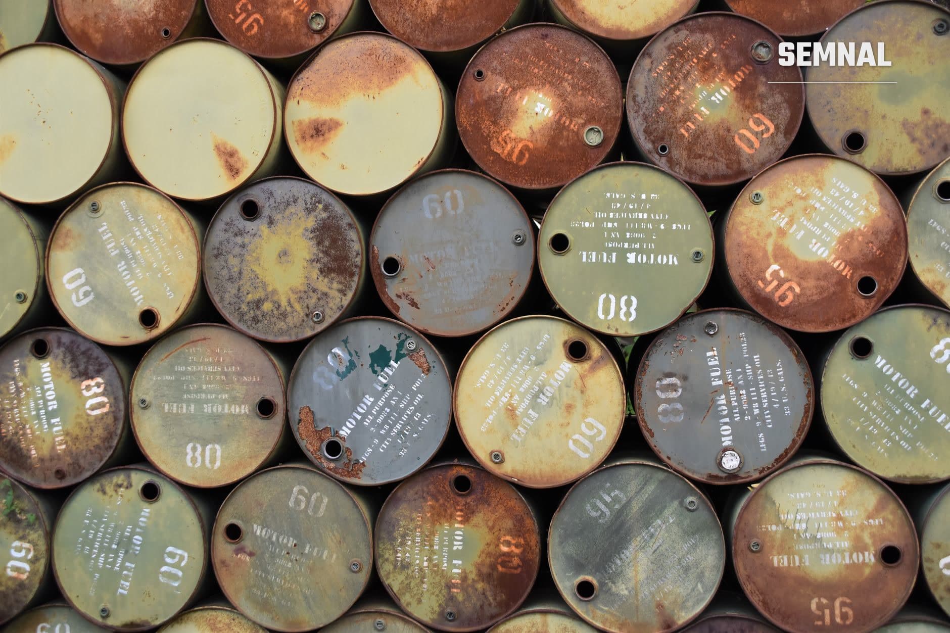Vedere de sus a mai multor bidoane metalice de combustibil deteriorate de rugină, cu numerotare și marcaje gravate pe capacele oxidate, stivuite în depozitare.