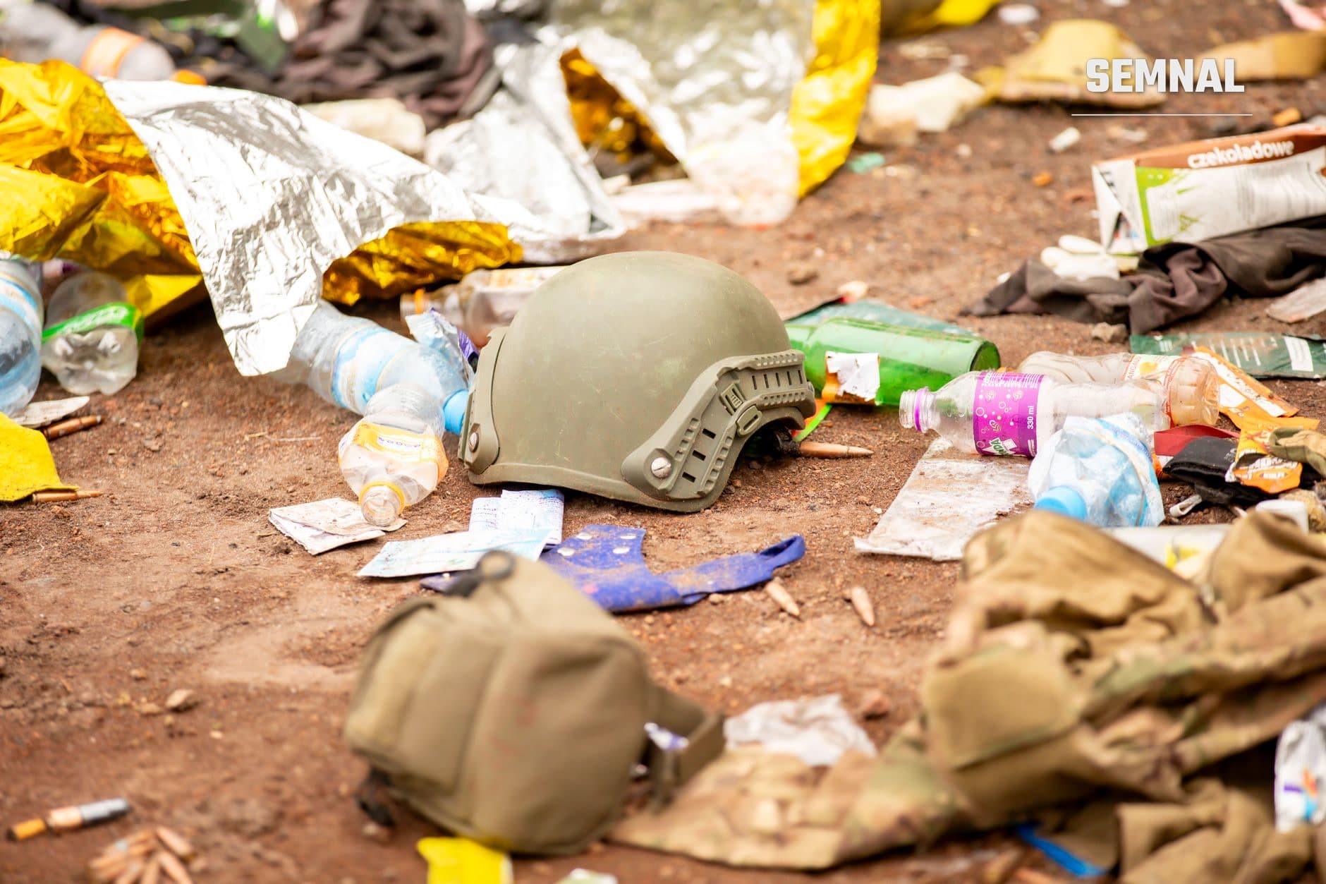 Cască militară gri-bej și echipament militar degradat împreună cu resturi personale și ambalaje pe teren gol aridos, dovezi ale unui conflict armat în Sudan.