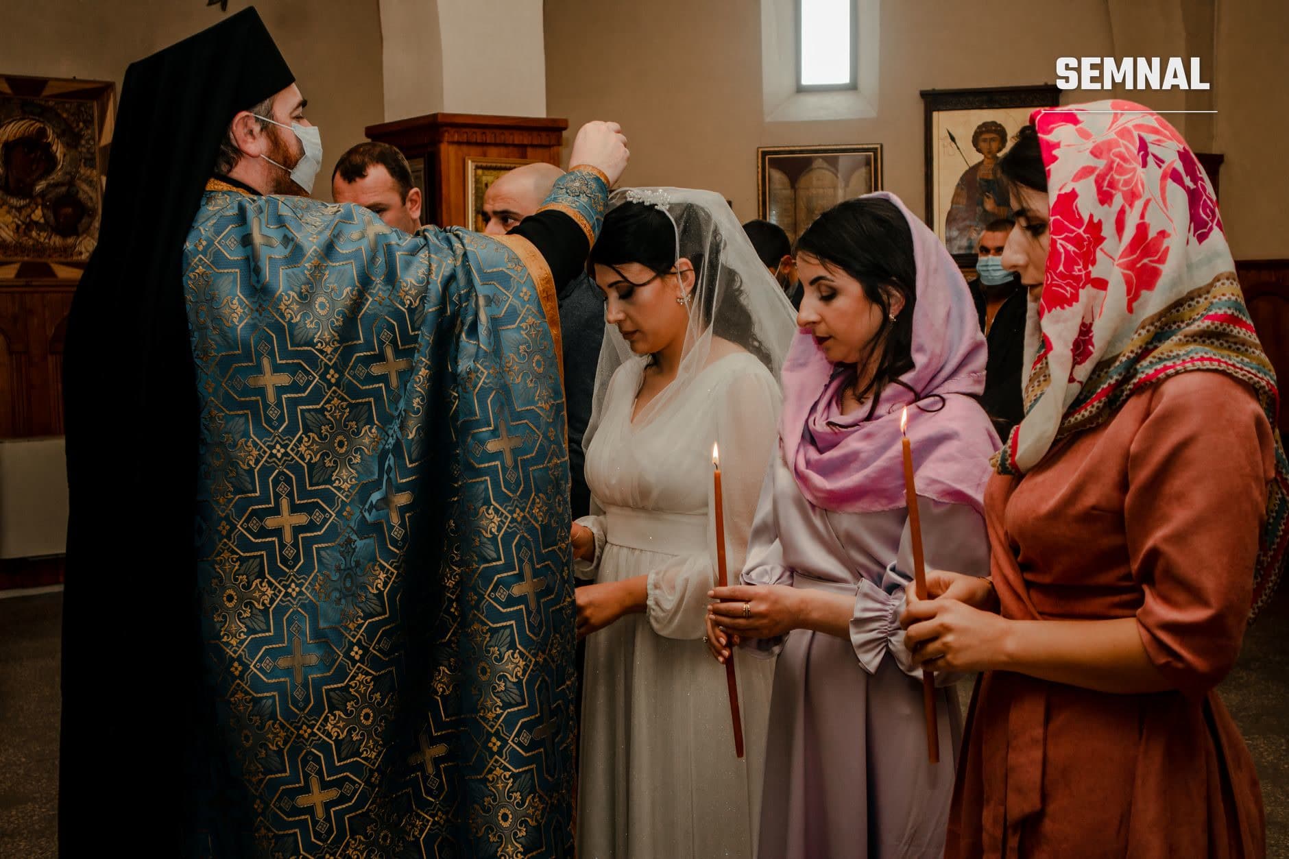 Preot ortodox cu vestminte albastru-aurii oferind binecuvântare unor femei credincioase cu capul acoperit în interiorul unei biserici, ilustrând tradiții și practici religioase românești contemporane.