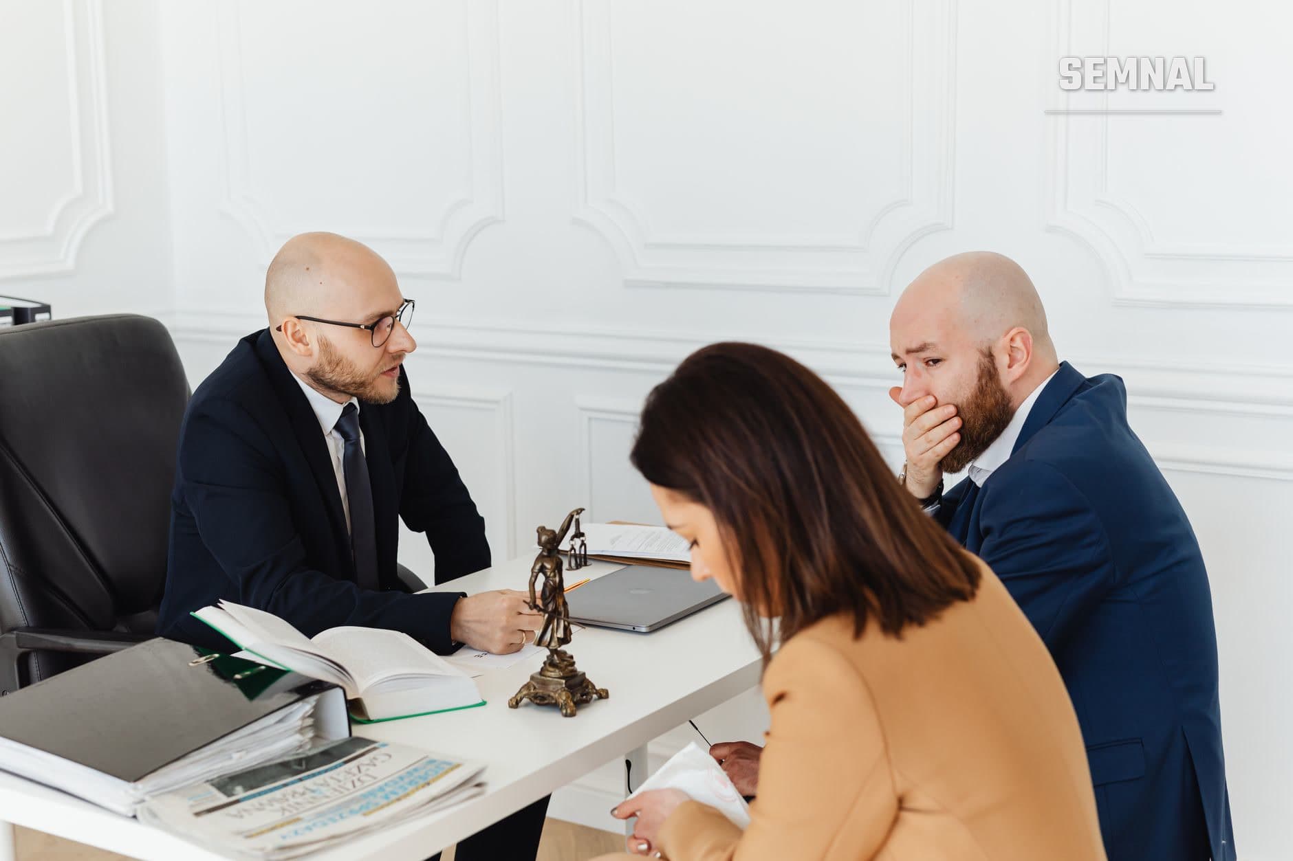 Trei persoane într-o birou juridic discută la masă cu documente și statueta Justiției, sugerând o sesiune de mediere sau consultație în caz de divorț.