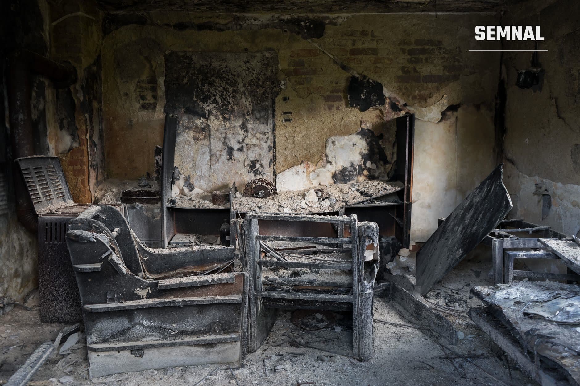 Interior al unei clădiri distruse de incendiu, cu pereți negriți, paturi metalice arse și resturi pe podea, ilustrând urmele unui dezastru tragic.