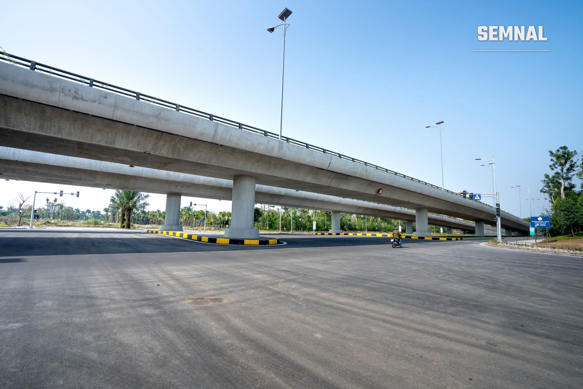 Autostradă modernă cu viaduct din beton și marcare galben-neagră, iluminare solară și semn rutier albastru pe o cărări goală sub cer senin, ilustrând infrastructura de transport rutier actuală.