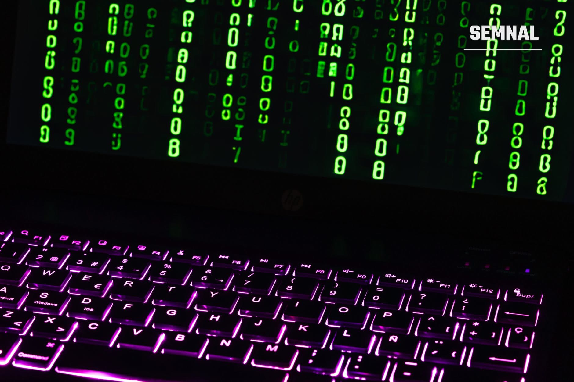 Tastatură cu iluminare neon mov și șiruri de cod binar verde curgând pe ecran într-o scenă cibernetică de hacker, pe fundal negru, reprezentând atacuri digitale.