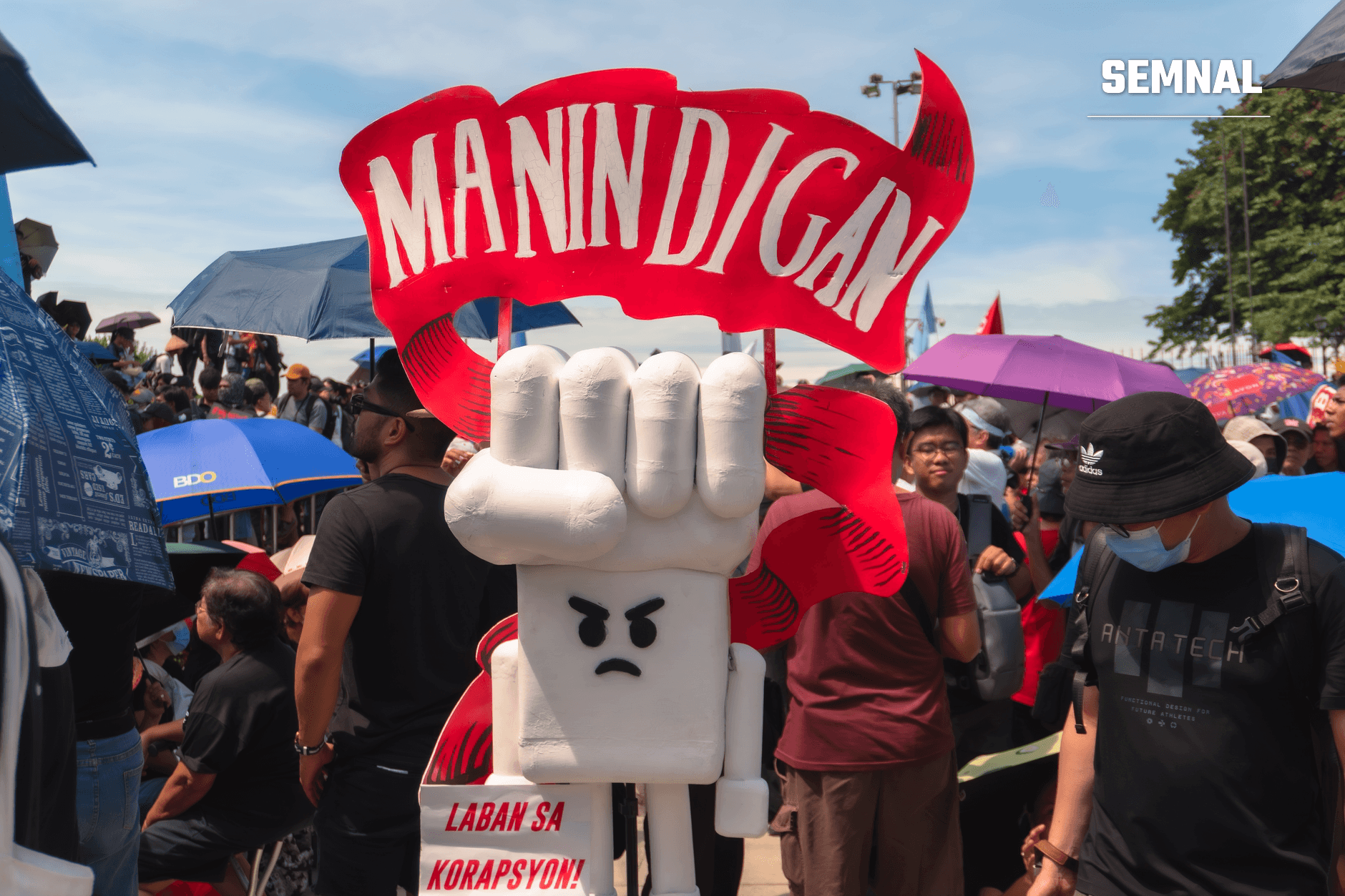 Mascot cu pumn ridicat și expresie furioasă purtând banderolă roșie, în mijlocul unei manifestații împotriva corupției la Jakarta, cu mulțimea de protestatari în fundal.