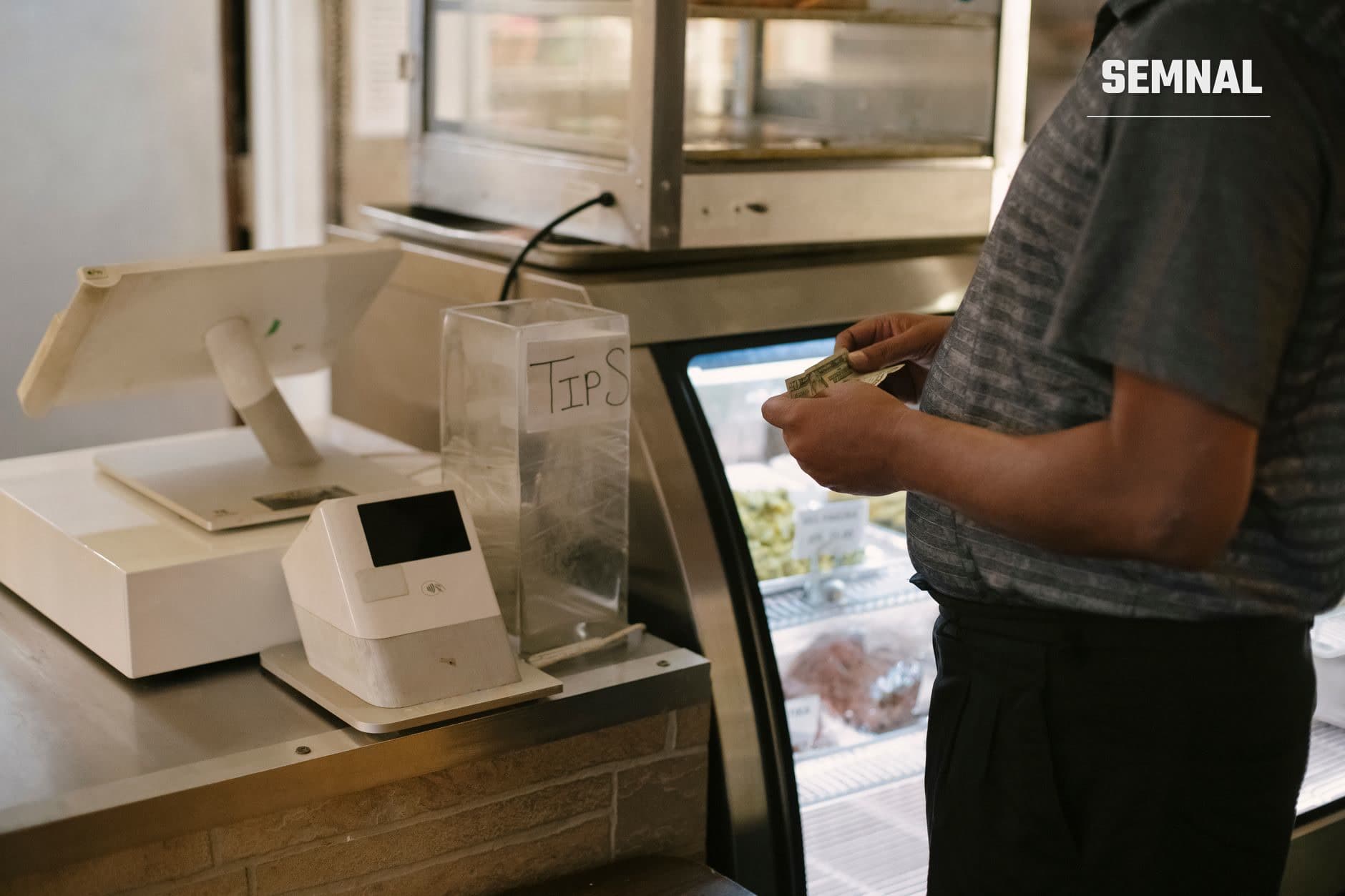 Persoană care introduce o bancnotă într-un recipient transparent etichetat 'TIPS' pentru bacșiș, într-un mediu de servicii cu echipamente de vânzare și frigider în fundal.