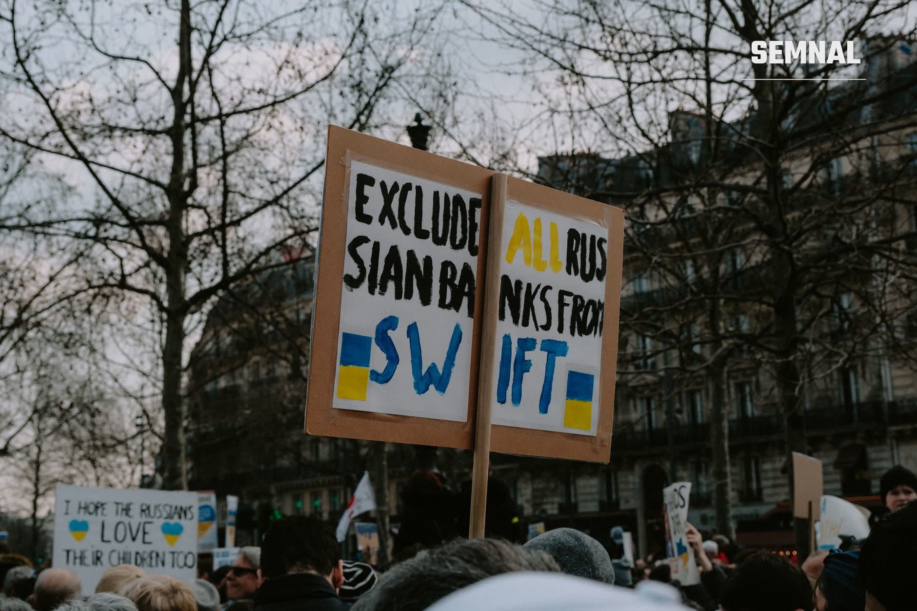 Manifestație publică cu participanți ținând pancarte manuscrise cu mesaje pro-Ucraina și anti-Rusia, inclusiv cereri de sancțiuni, în piață urbană cu copaci dezgoliți de frunze.