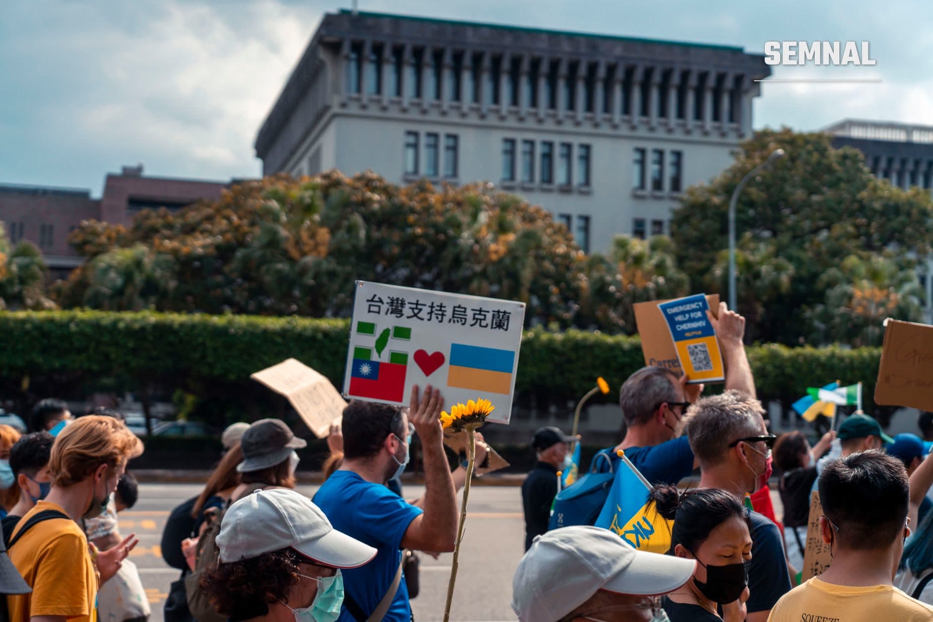 Manifestație de stradă cu suporteri ai Taiwanului ținând pancarte cu steaguri taiwanese și ucrainene, flori galbene și mesaje de solidaritate în fața unui edifciu administrativ modern.