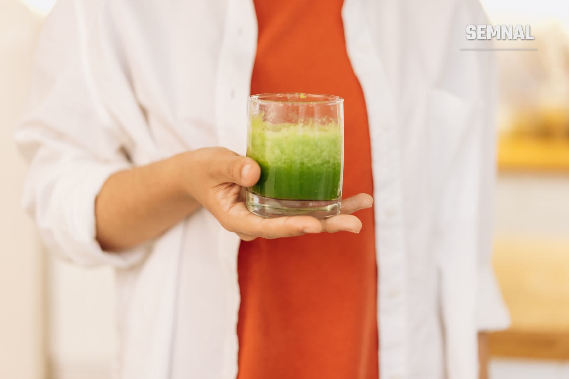Persoană ținând un pahar cu suc verde vibrant, purtând cămașă albă și vestă roșie, ilustrând curele de detoxifiere cu sucuri.