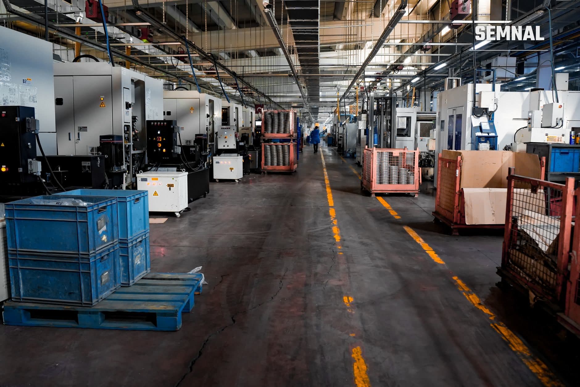 Hală de producție industrială cu mașini CNC, containere de transport și muncitori pe podea marcată cu liniatura galbenă.