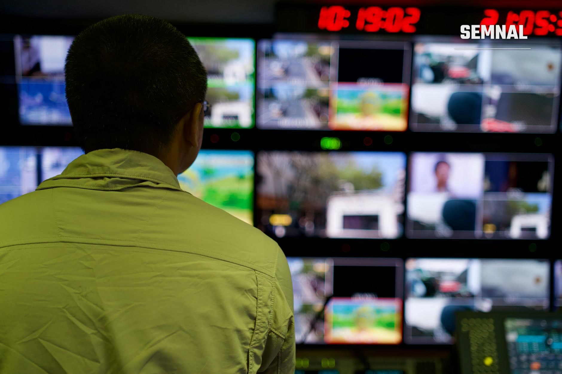 Operator de supraveghere privind un panou cu opt ecrane de monitoring color care afișează diverse locații în timp real, vorbind cu o jachetă galbă, în centru de control cu indicatori luminoși verzi.