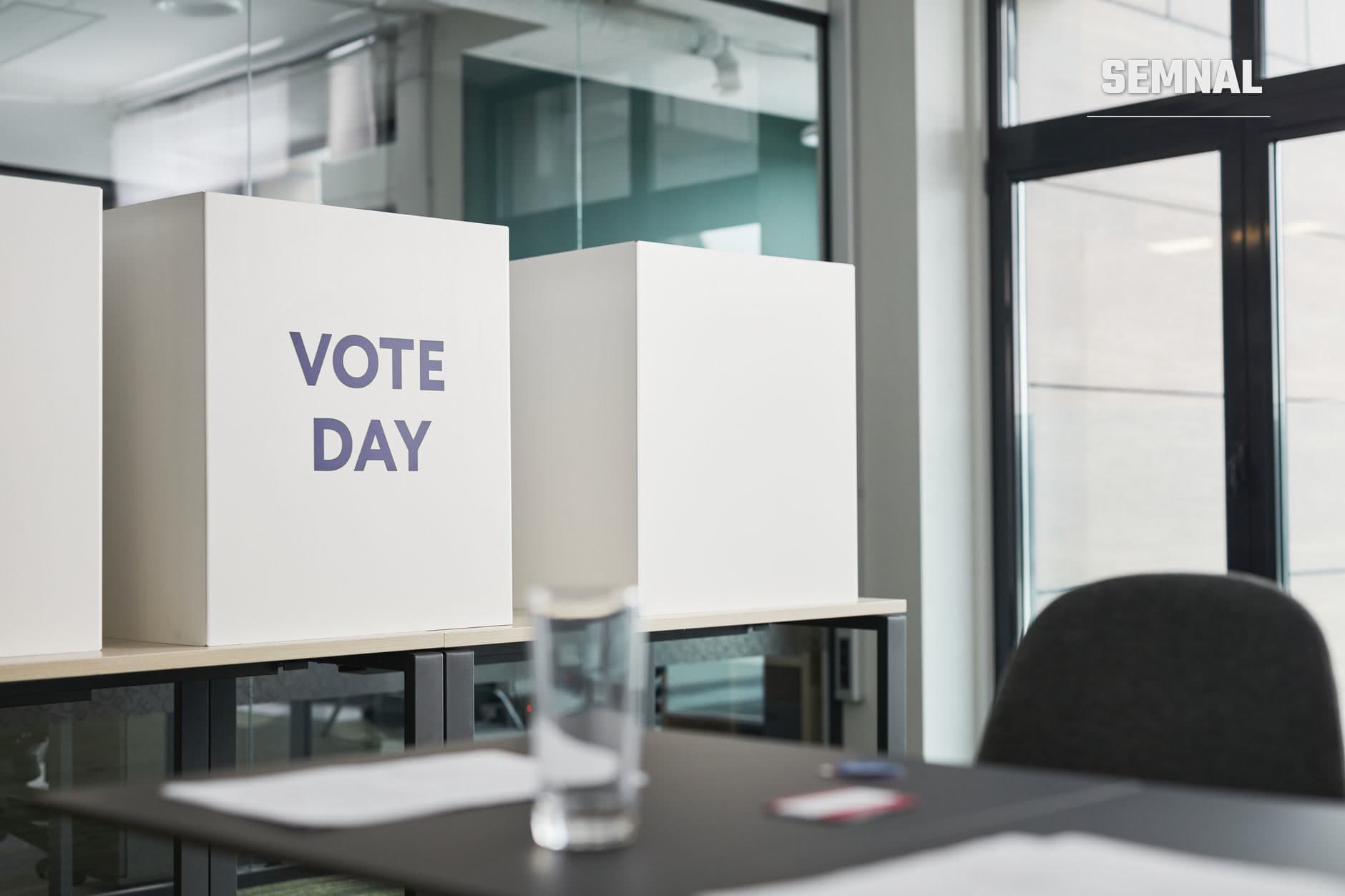 Cutii de vot albe cu inscripția 'VOTE DAY' amplasate pe masă din birou modern, sugerând proces electoral și participarea civică la o rundă de scrutin.