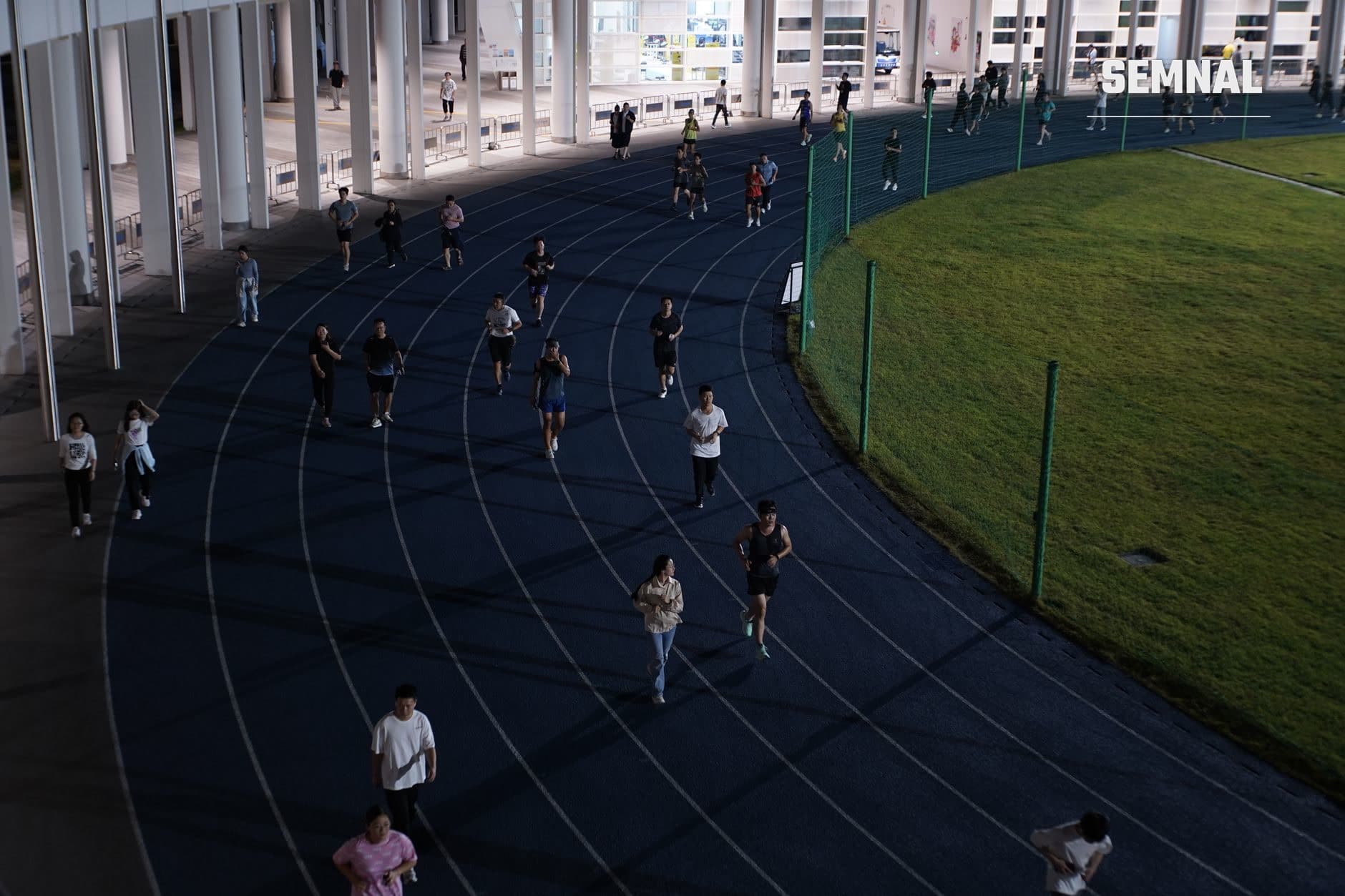 Zeci de alergători distribuiți pe pista de atletism curbă, sub structură de beton, alergând și antrenând în grup pe benzi paralele marcate, evidențiind comunitatea și cursele populare