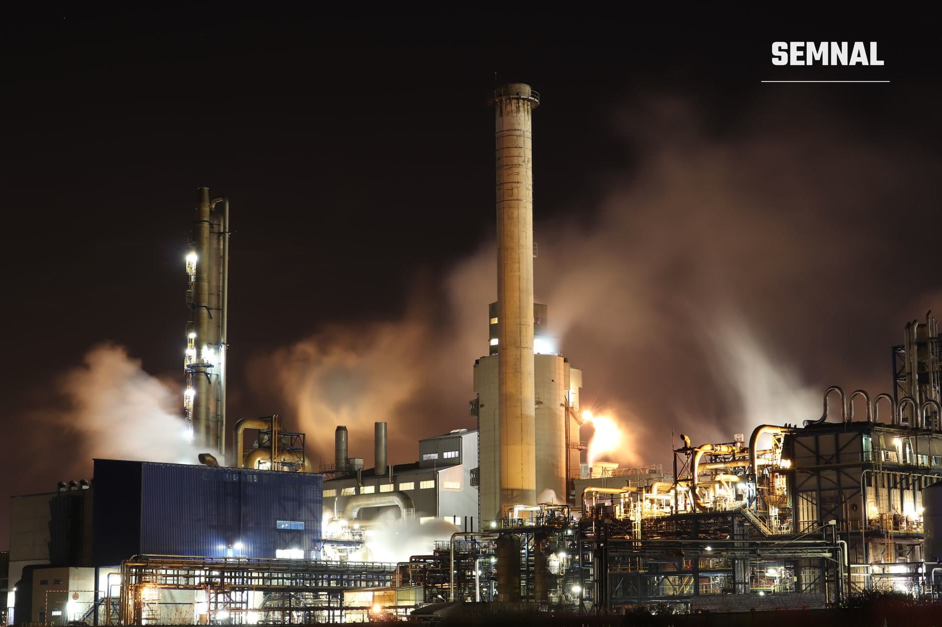 Rafinărie de petrol noaptea cu turnuri de distilare iluminate, fum dens și flăcări de incendiu din bombardament, structuri industriale iluminate de explozii și foc aprins la o instalație petrolieră.