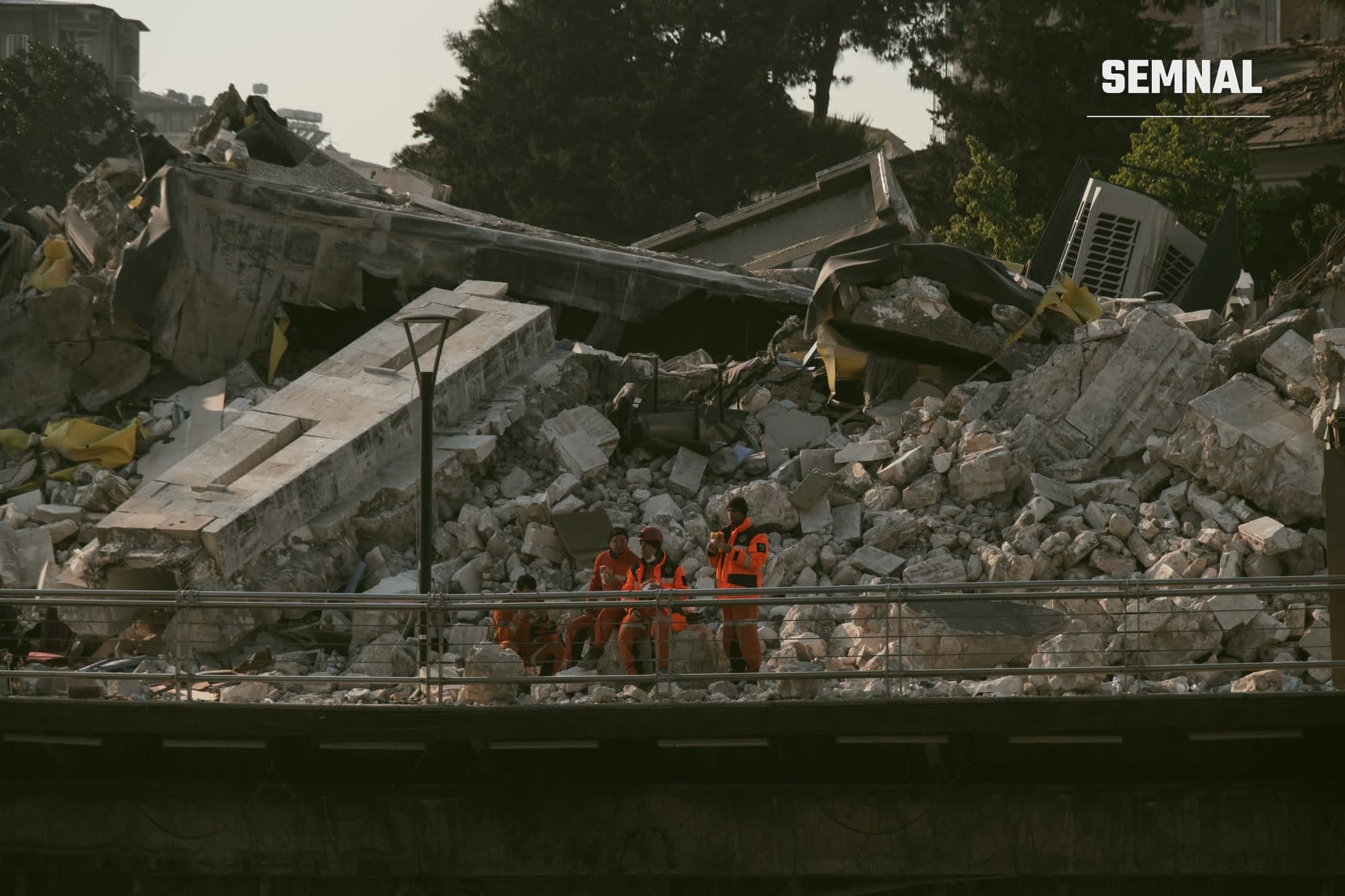 Echipă de muncitori în costume de protecție portocalii lucrând în mijlocul dărâmăturilor unei clădiri prăbușite, cu excavatoare galbene și resturi masive de beton pe pământ.