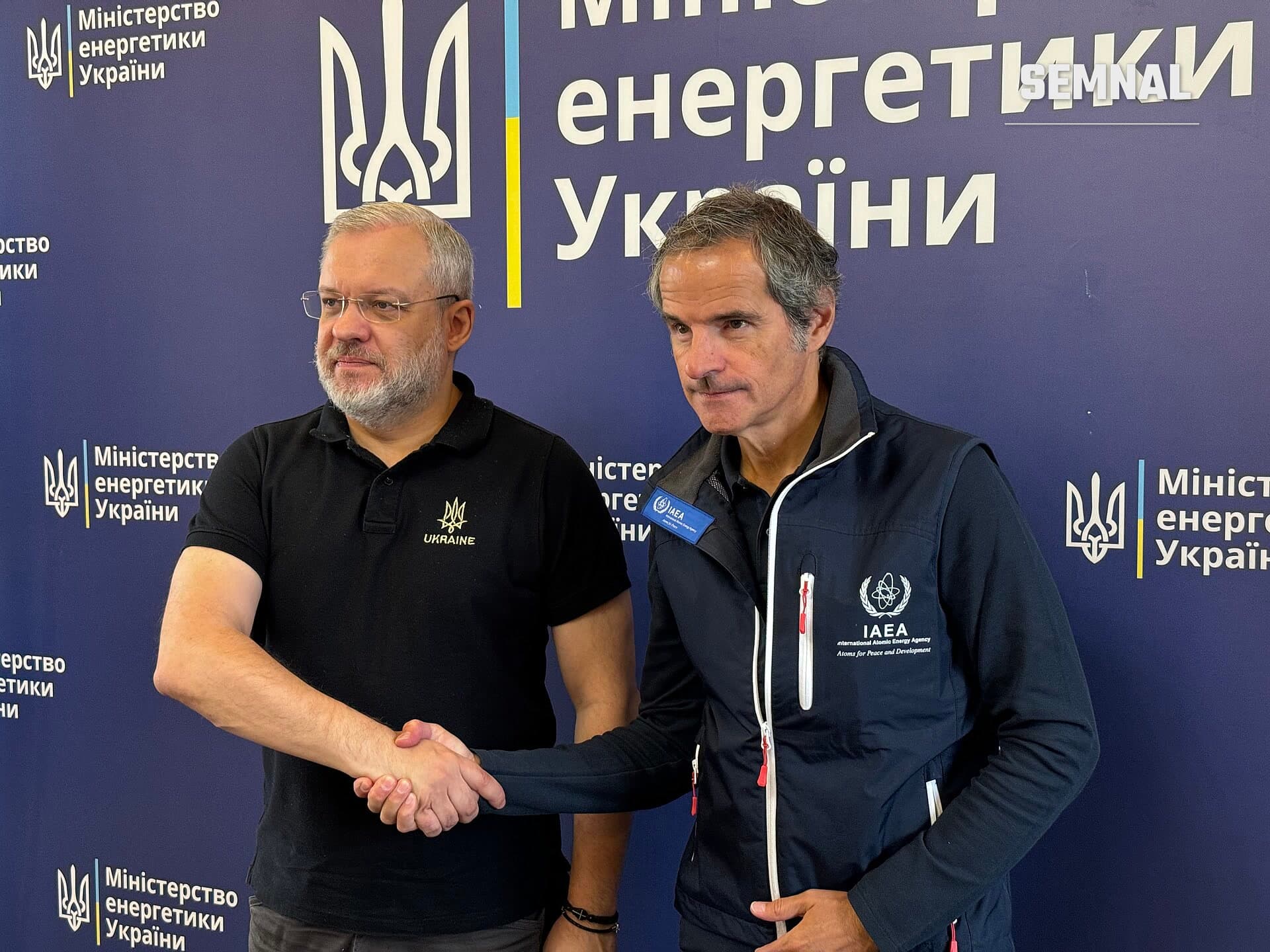 Rafael Grossi, director general al AIEA, se dă mâna cu un oficial din Ministerul Energeticii Ucrainei la o conferință oficială la Kiev, în fața unui banner institucional. (Foto: IAEA Imagebank)