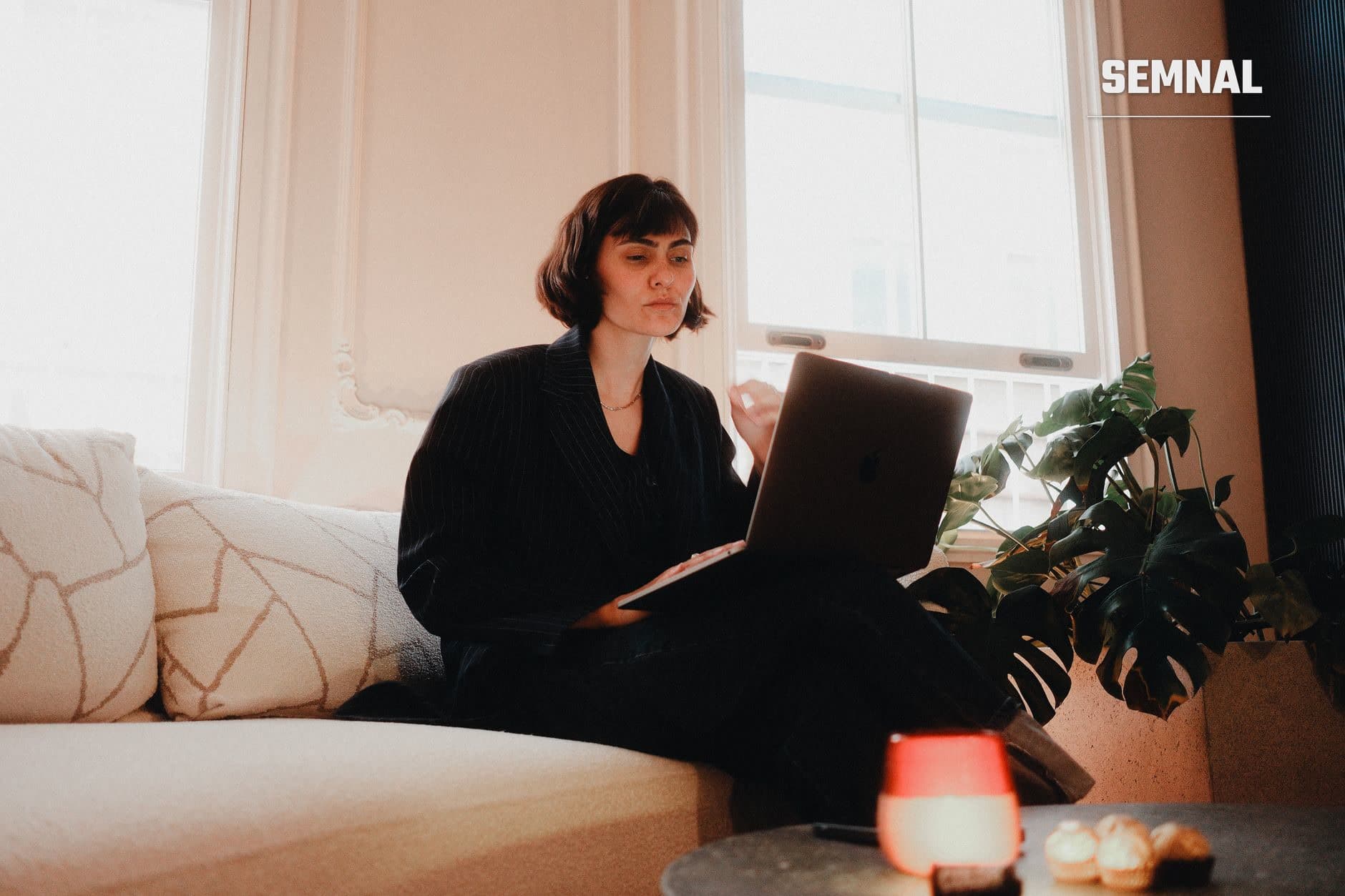 Femeie care lucrează de acasă pe laptop, ședând pe canapea lângă fereastră, cu plantă decorativă și cană pe masă (Foto: Mert Coșkun / Pexels)