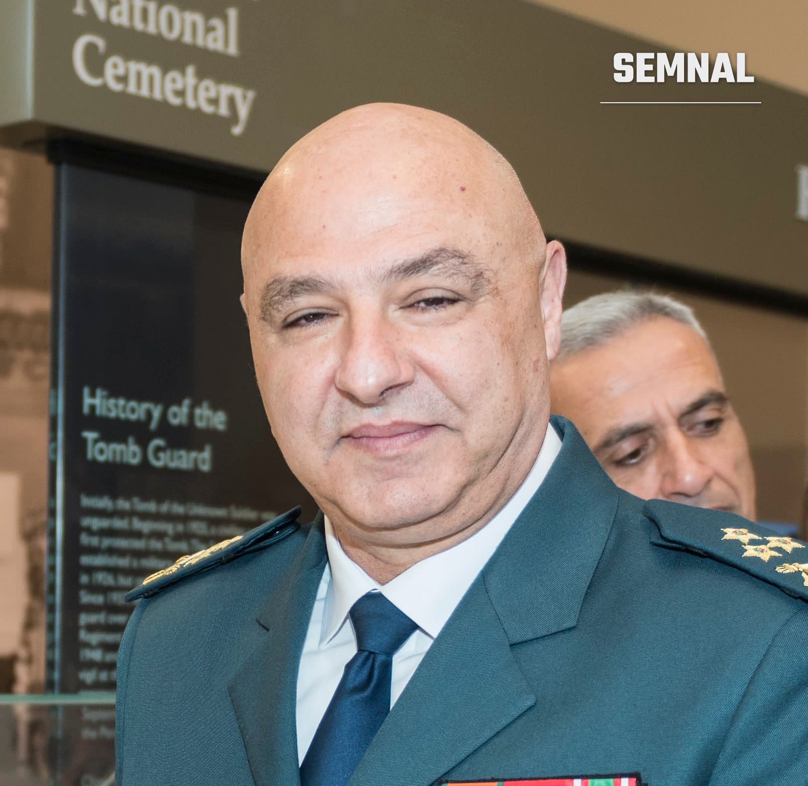 Joseph Aoun, președintele Libanului, purtând uniformă militară gri cu decorații și eșarfă albastră, fotografiat la o instituție oficială cu panou informativ în fundal. (Foto: Arlington National Cemetery)