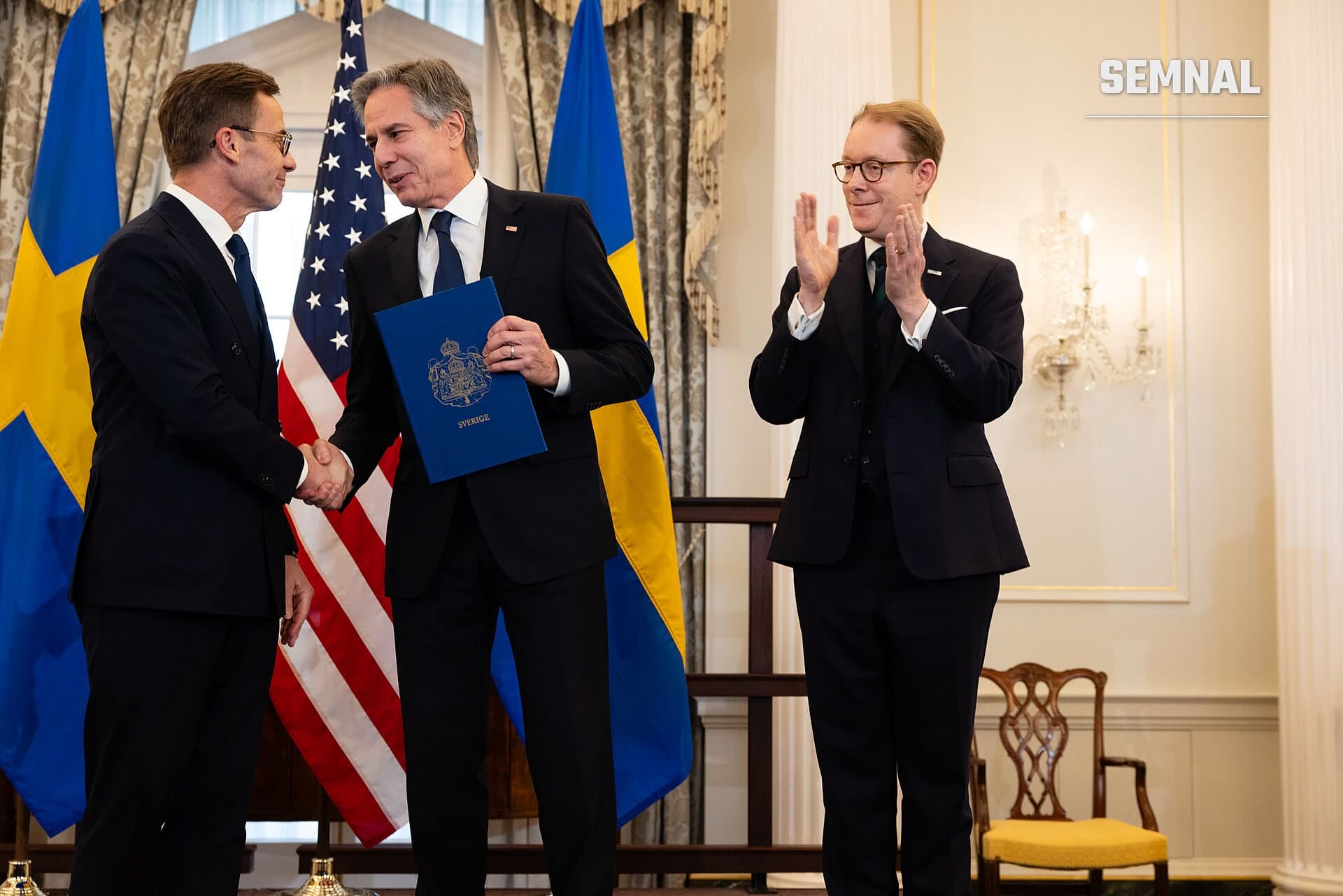 Ulf Kristersson, premierul Suediei, participând la o ceremonie oficială în costum negru, ținând un document cu stema Suediei, alături de doi oficiali, cu drapelele Suediei și Statelor Unite în fundal. (Foto: U.S. Department of State)