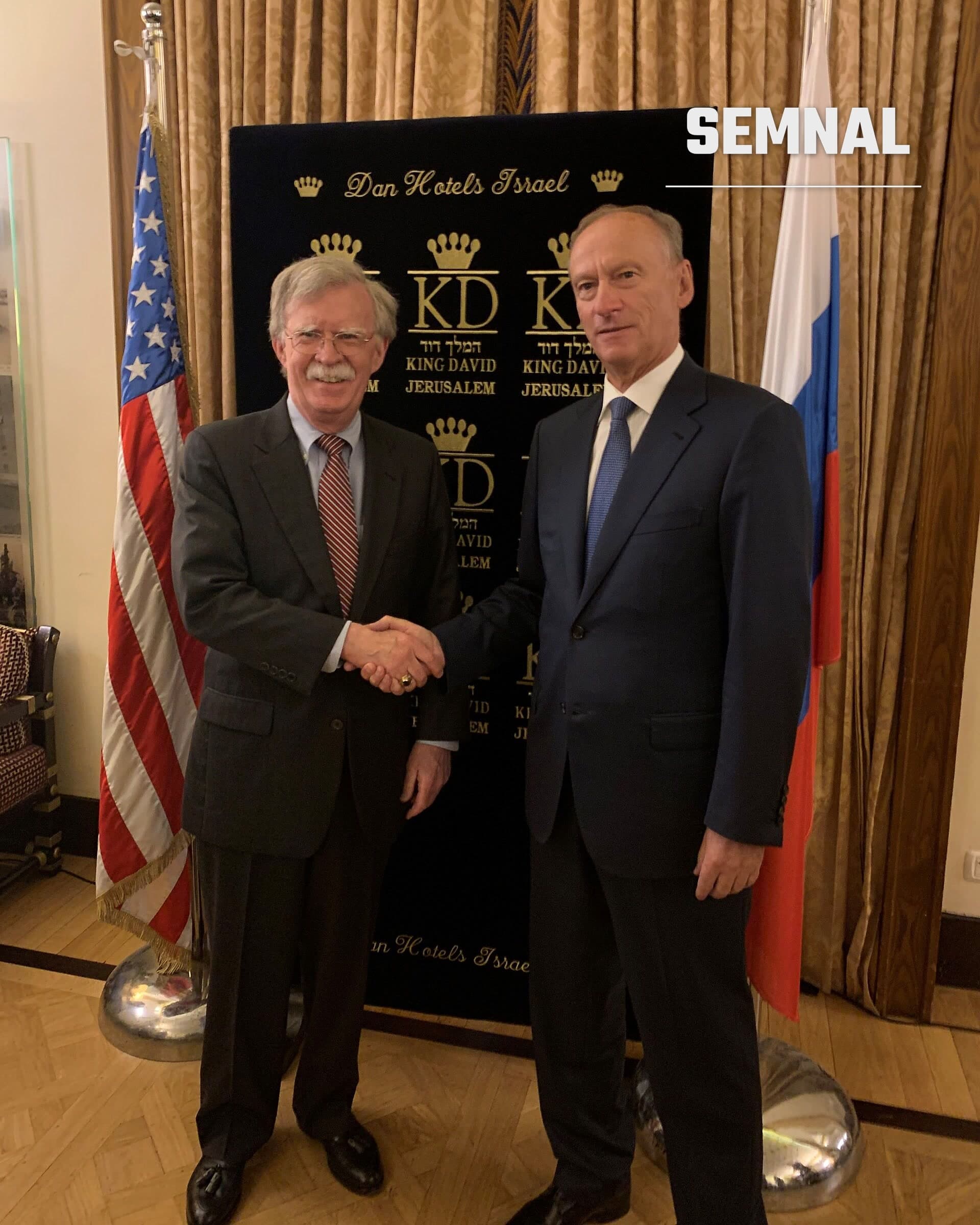 Nikolai Patrushev în costum negru și cravată albastră dând mâna cu un oficial american, în fața drapelelor SUA și Rusiei, la o întâlnire oficială la King David Jerusalem Hotel. (Foto: Office of U.S. National Security Advisor)