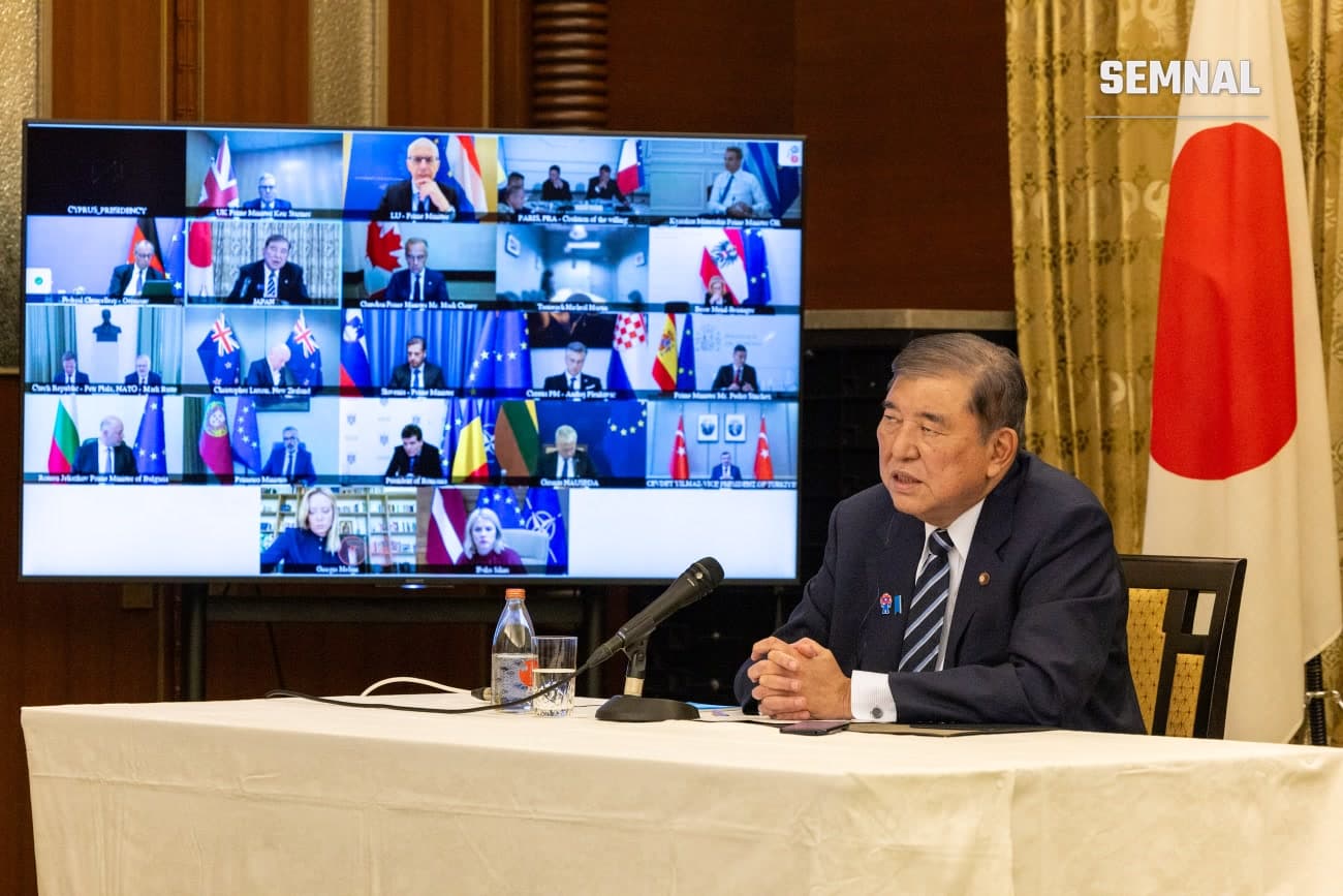 Nicușor Dan, prezident, la o conferință sau consultări oficiale, șezând la masă cu microfon în fața unui ecran cu participanți internaționali, în fața steagului Japoniei. (Foto: Cabinet Public Affairs Office of Japan)