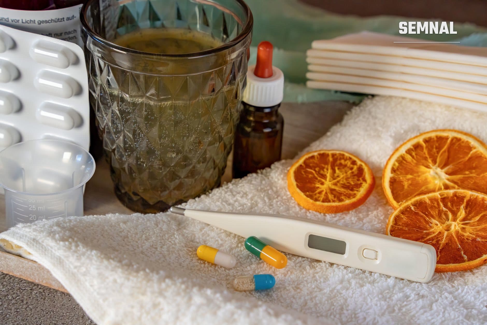 Tocuri medicale și remedii naturale pentru răceală: pahar cu ceai, portocale tăiate, termometru digital, comprimate colorate și sticlă de sirop pe prosop alb (Foto: Gundula Vogel / Pexels)