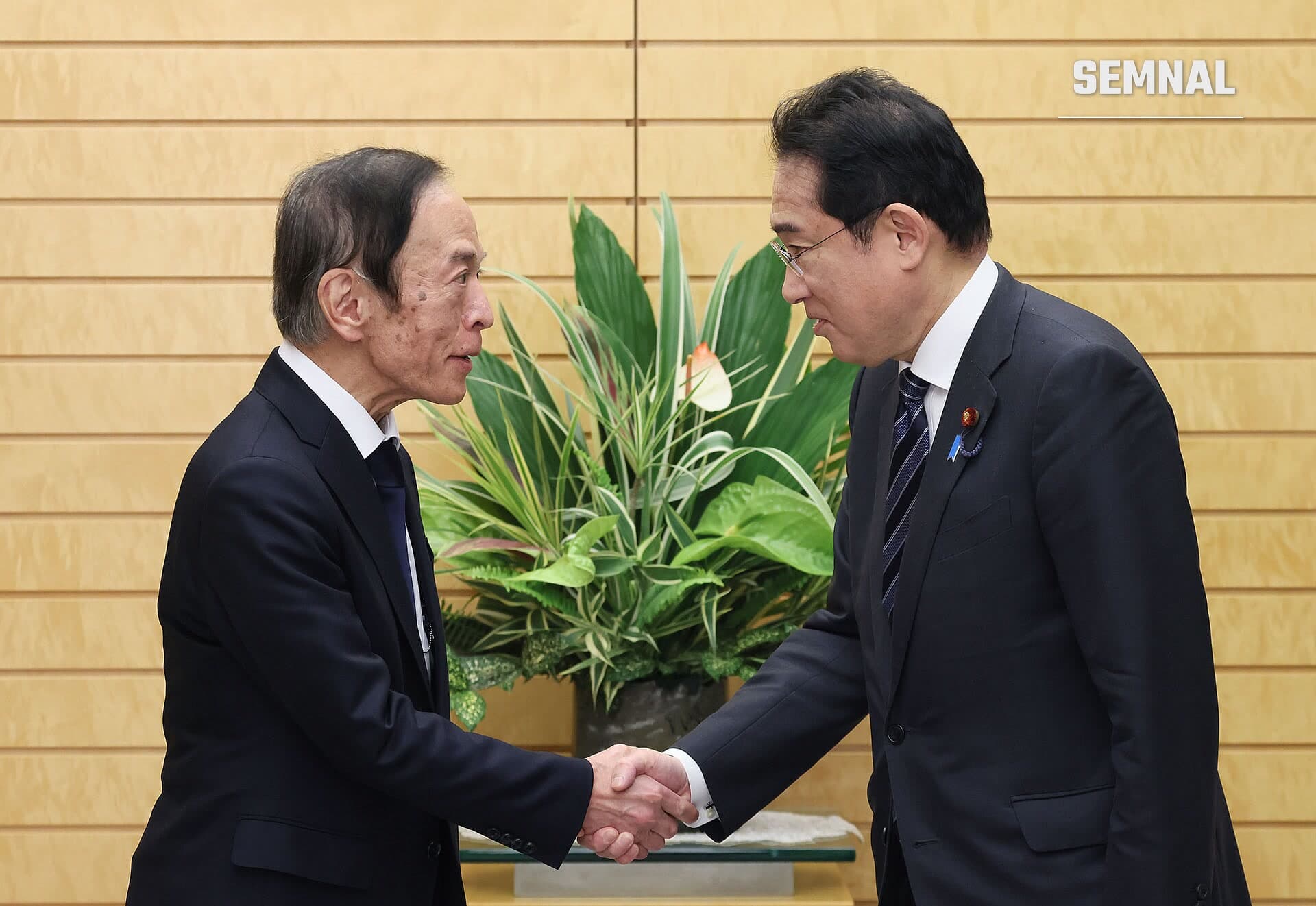 Kazuo Ueda, guvernatorul Băncii Japoniei, strânge mâna cu un oficial în costum negru la o reuniune formală, cu o plantă verde în fundal. (Foto: 内閣官房内閣広報室)