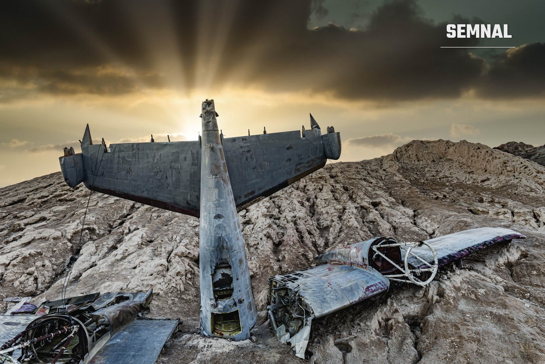 Epavă de avion militar cu fuselaj sfâșiat și aripi zdrobite, răspândite pe teren rocoso și nisipou într-un peisaj desert sub cer purtător de nori grei și raze solare dramatice. (Foto: Michael Gattorna / Pexels)