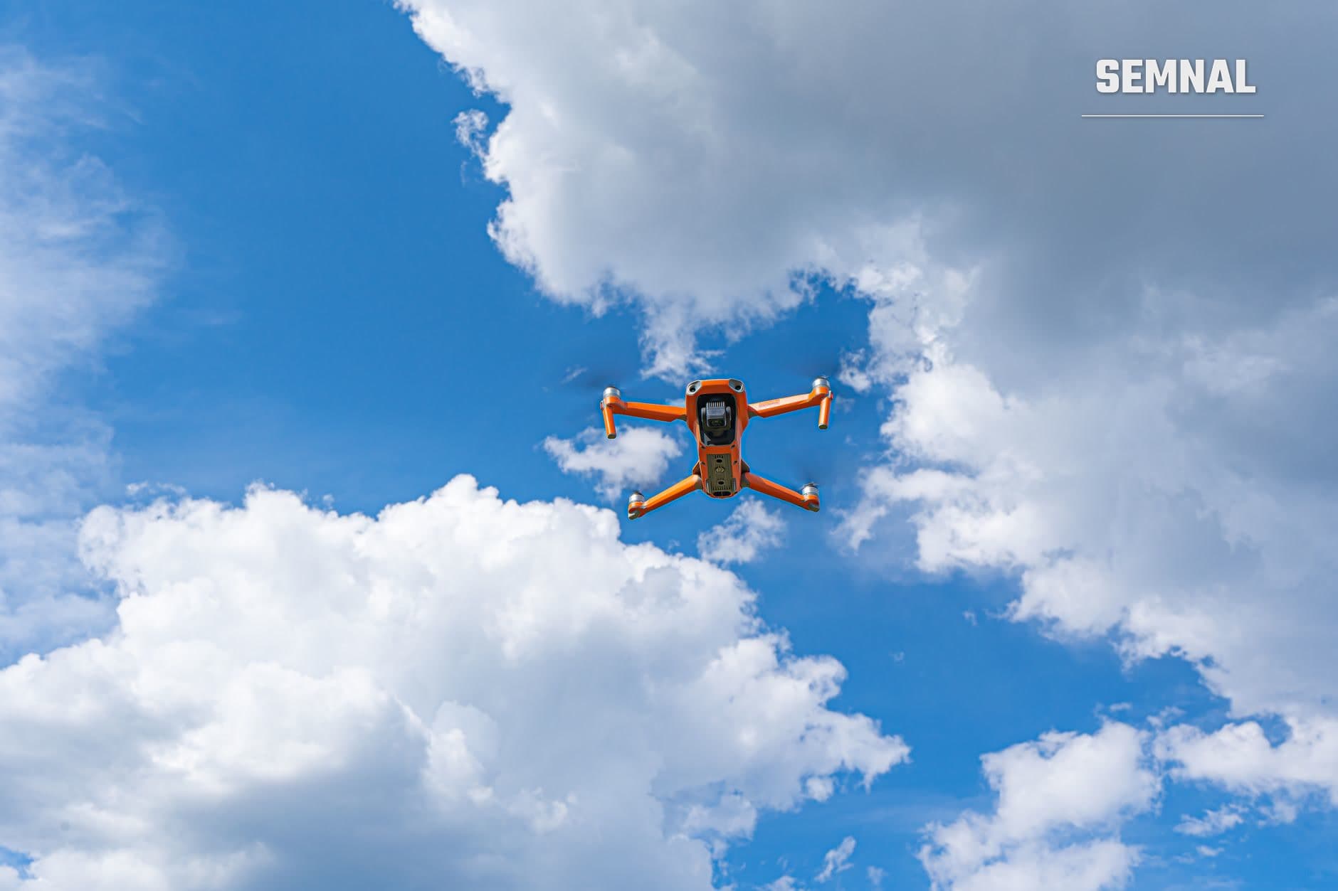 Dronă portocalie cu patru elice zburând pe cer albastru cu nori albi, fotografiată de jos în sus în poziție de zbor. (Foto: Castorly Stock / Pexels)