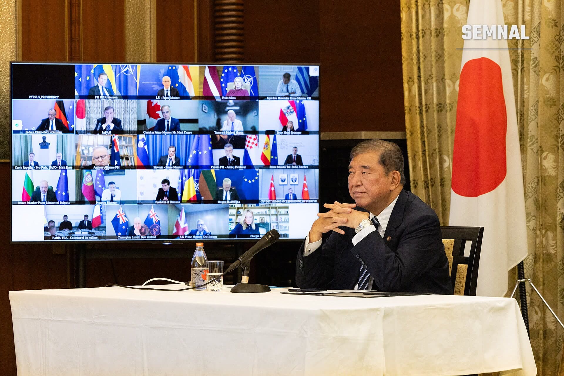 Nicușor Dan, în costum negru, vorbind la o conferință video internațională cu mai mulți lideri europeni și internaționali pe ecran, steag roșu și alb al Japoniei în fundal (Foto: Cabinet Public Affairs Office of Japan)