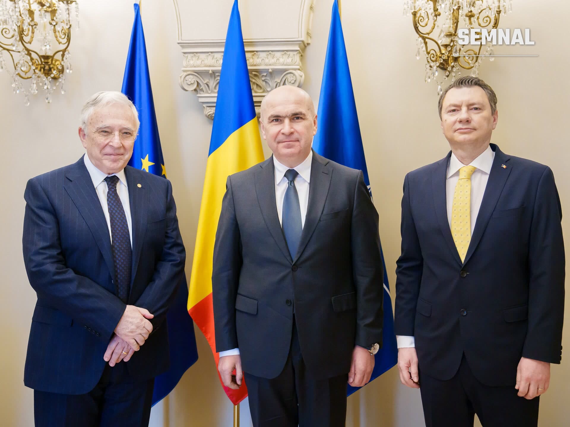 Ilie Bolojan, prim-ministrul României, în costum gri și cravată albastră, pozat oficial în fața steagurilor României și Uniunii Europene, alături de doi colegi în costume de afaceri. (Foto: Presidential Administration of Romania)