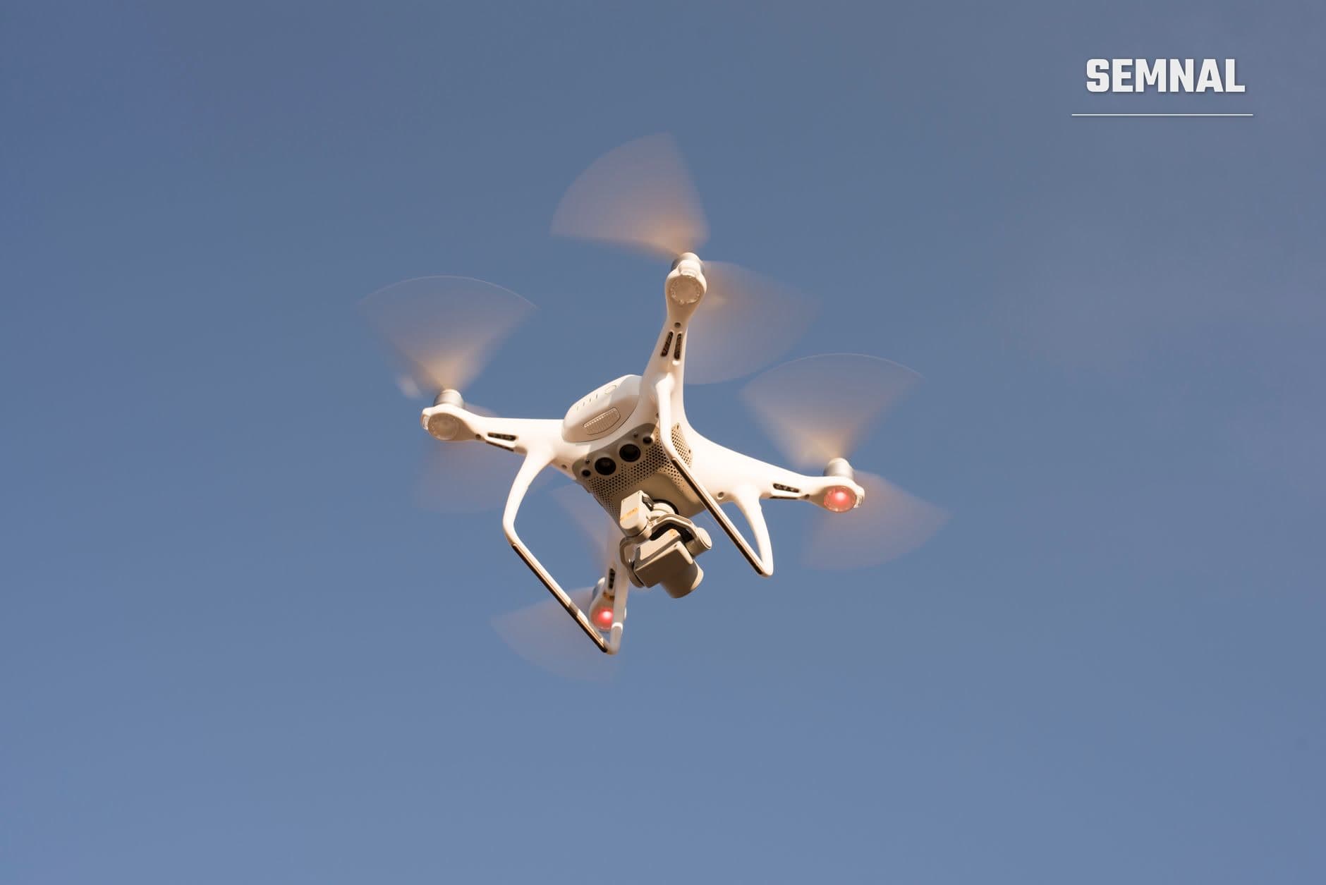 Dronă quadcopter albă în zbor pe cer albastru, cu patru elice rotitoare și cameră suspendată, echipată cu indicatoare luminoase roșii de poziție (Foto: Daniel Reche / Pexels)