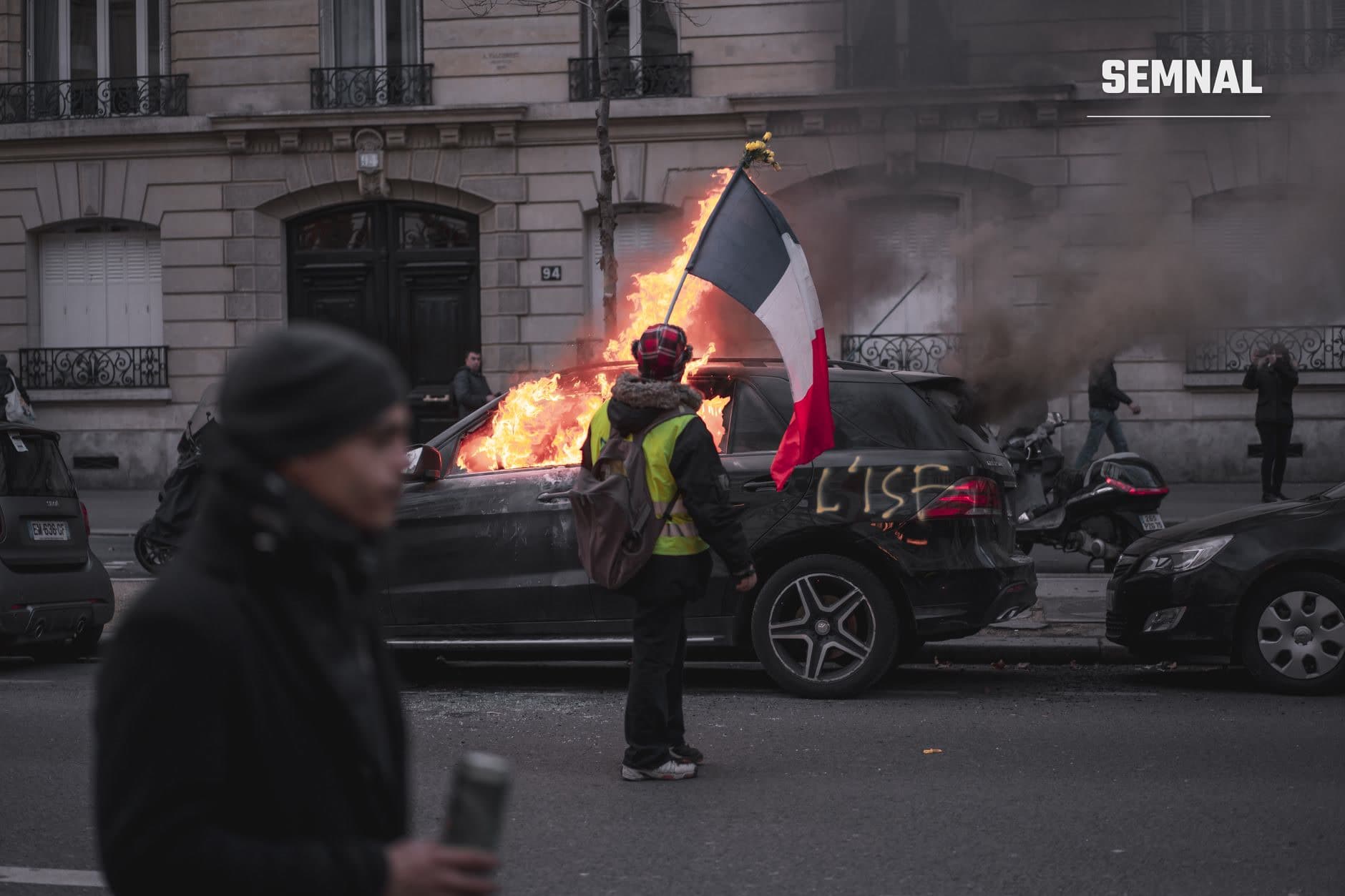 Manifestant cu vesta galbenă și steag francez în fața unei mașini în flăcări pe o stradă parizianță, în contextul protestelor violente din Franța. (Foto: Alotrobo / Pexels)