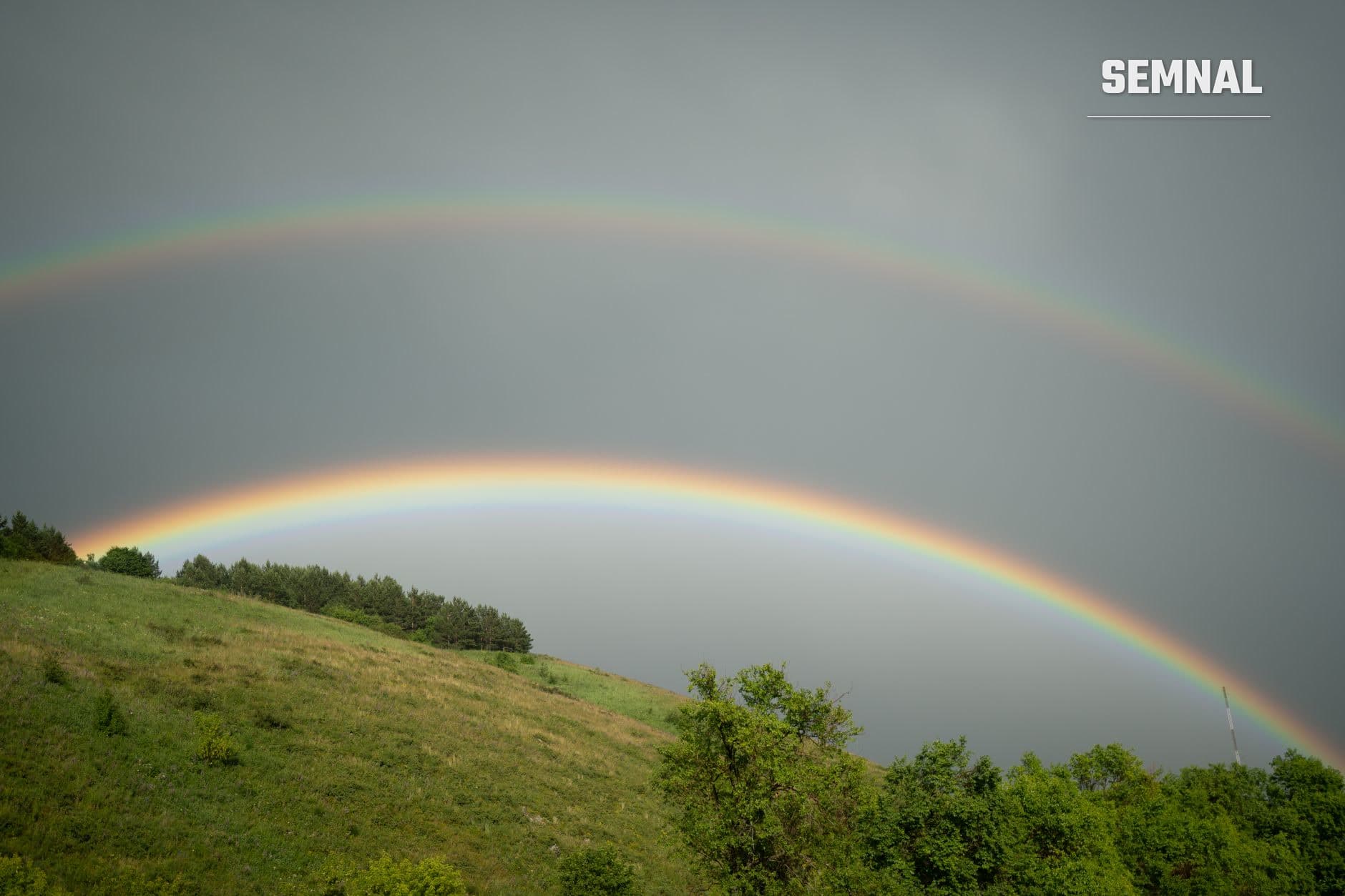 Curcubeu dublu spectaculos pe cer gri cu versant verde și pădure în prim-plan; curcubeul secundar arată ordinea culorilor inversată deasupra celui primar. (Foto: Dmitriy Piskarev / Pexels)