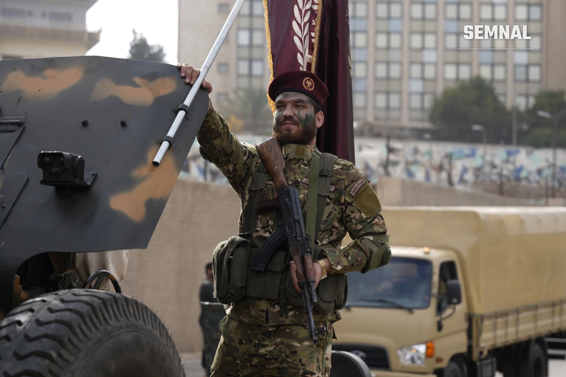 Militar în echipament tactic purtând uniform de camuflaj și pălărie tradițională, ținând o stindar militară bordeală și o armă, lângă vehicul militar într-o maniestație de forță în Orientul Mijlociu. (Foto: Baraa Obied / Pexels)