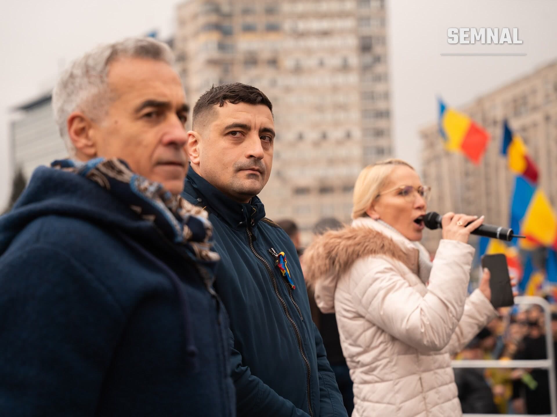 George Simion, liderul AUR, la o manifestație publică, purtând jachetă albastră, alături de alte persoane, cu steaguri ale României în fundal și clădiri ale unui oraș în spatele grupului. (Foto: AUR Alianța pentru Unirea Românilor)