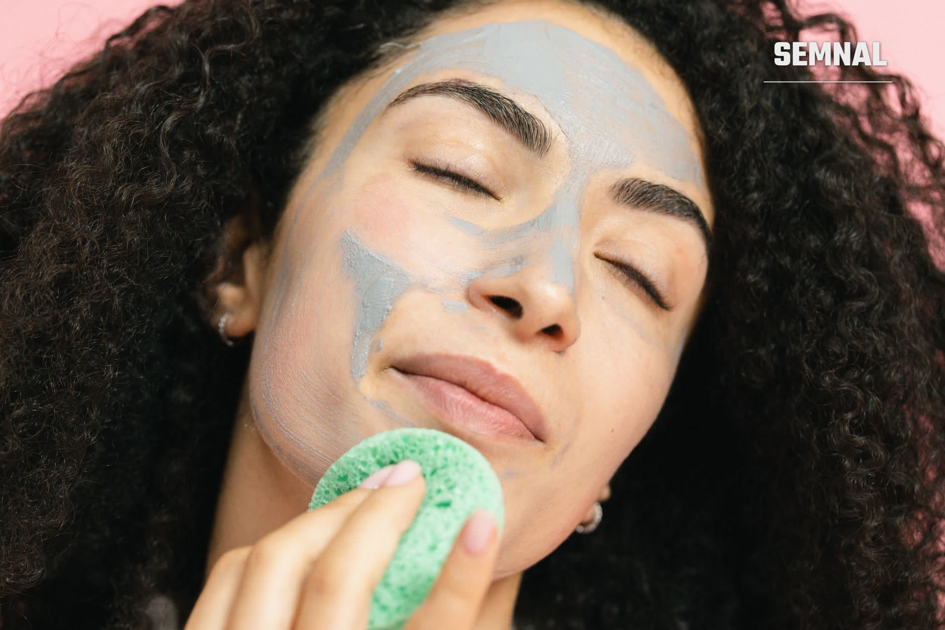 Femeie care aplică o mască de îngrijire a pielii albastru-gri pe față cu ajutorul unei bureți verzi, în cadrul unei rutine de frumusețe și ingrijire dermatologică. (Foto: SHVETS production / Pexels)
