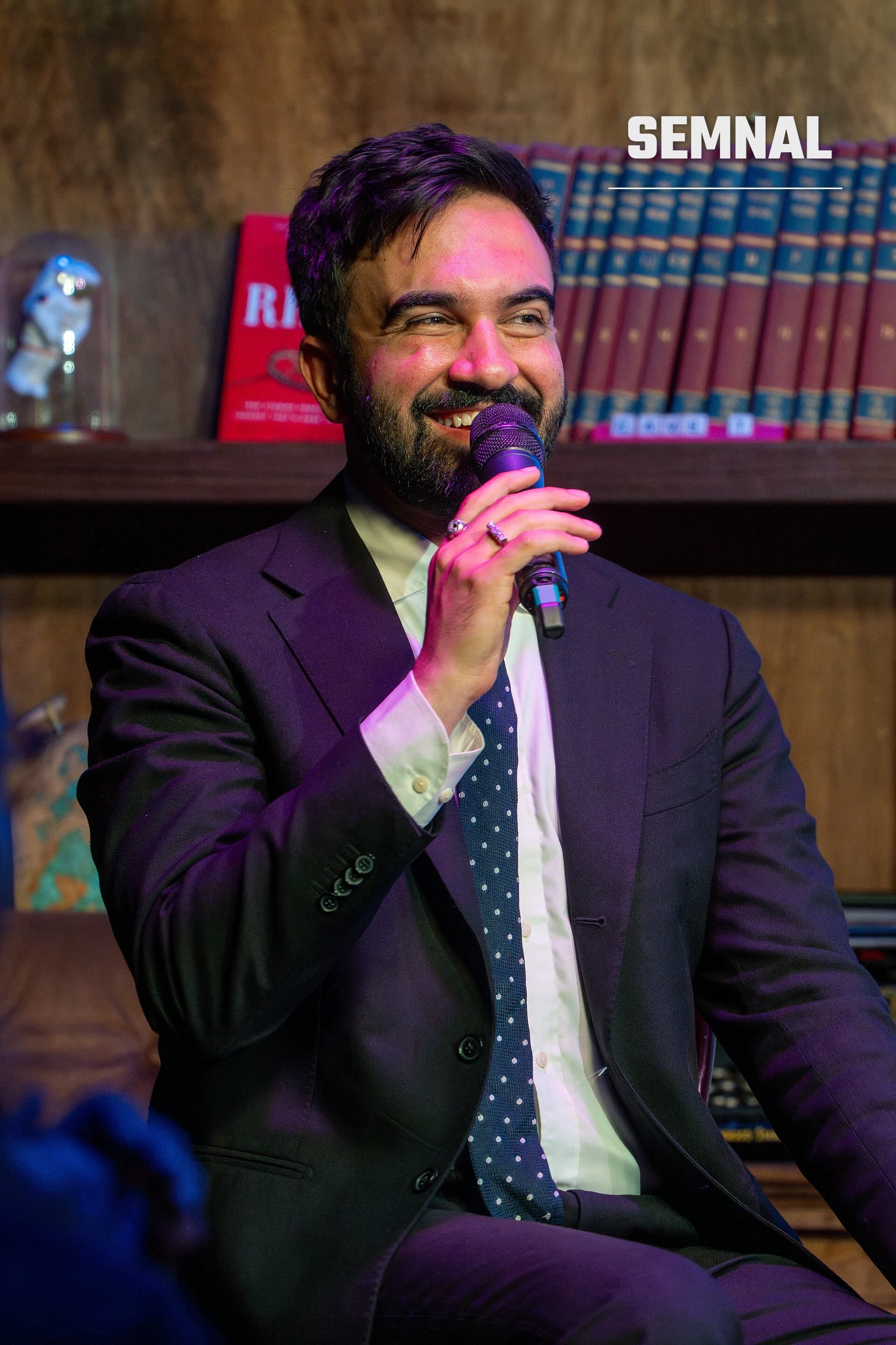 Zohran Mamdani, bărbat adult cu barbă, în costum bleumarin și cravată albastră, ținând un microfon la un eveniment public în interior, cu rafi de cărți în fundal și iluminare roz-albastră. (Foto: Bryan Berlin)