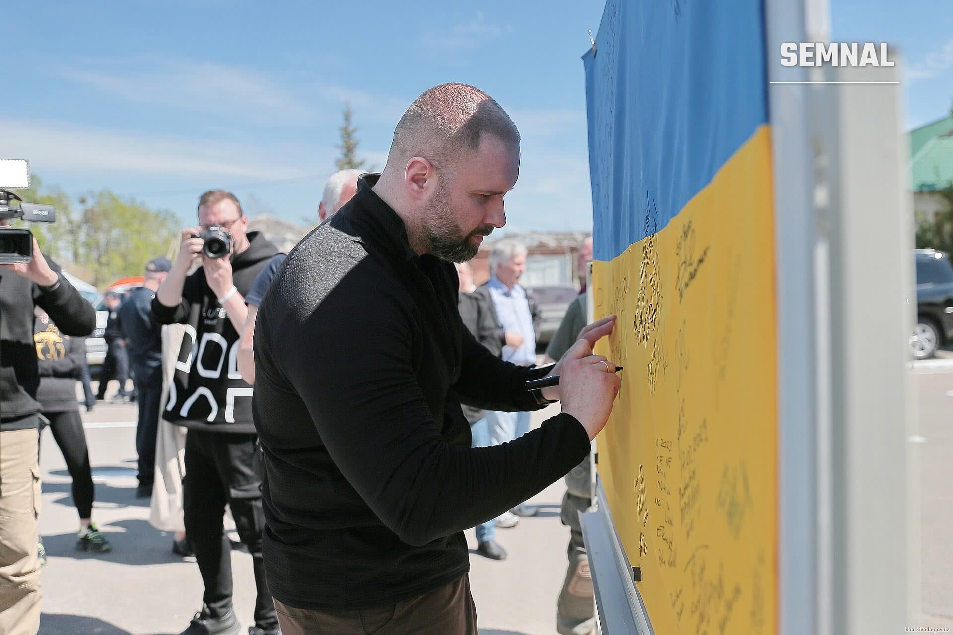 Oleh Syniehubov scrie pe o suprafață galbenă la un eveniment public, purtând jachetă neagră. (Foto: Харківська обласна військова адміністрація)