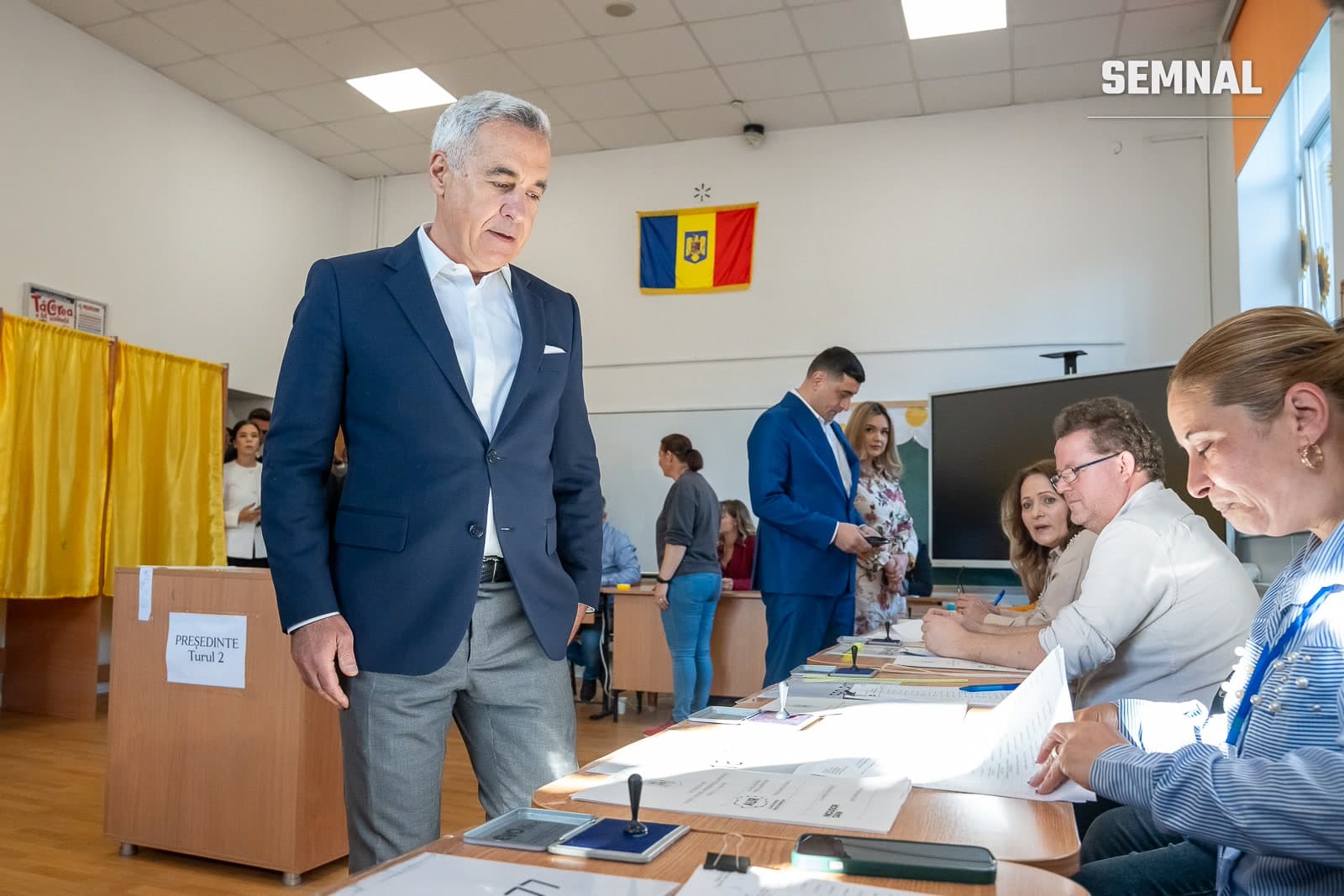 Călin Georgescu în costum bleumarin, cu cămașă albă și pantaloni gri, într-o secție de votare, privind spre masa cu ștampila și hârtiile. (Foto: AUR Alianța pentru Unirea Românilor)