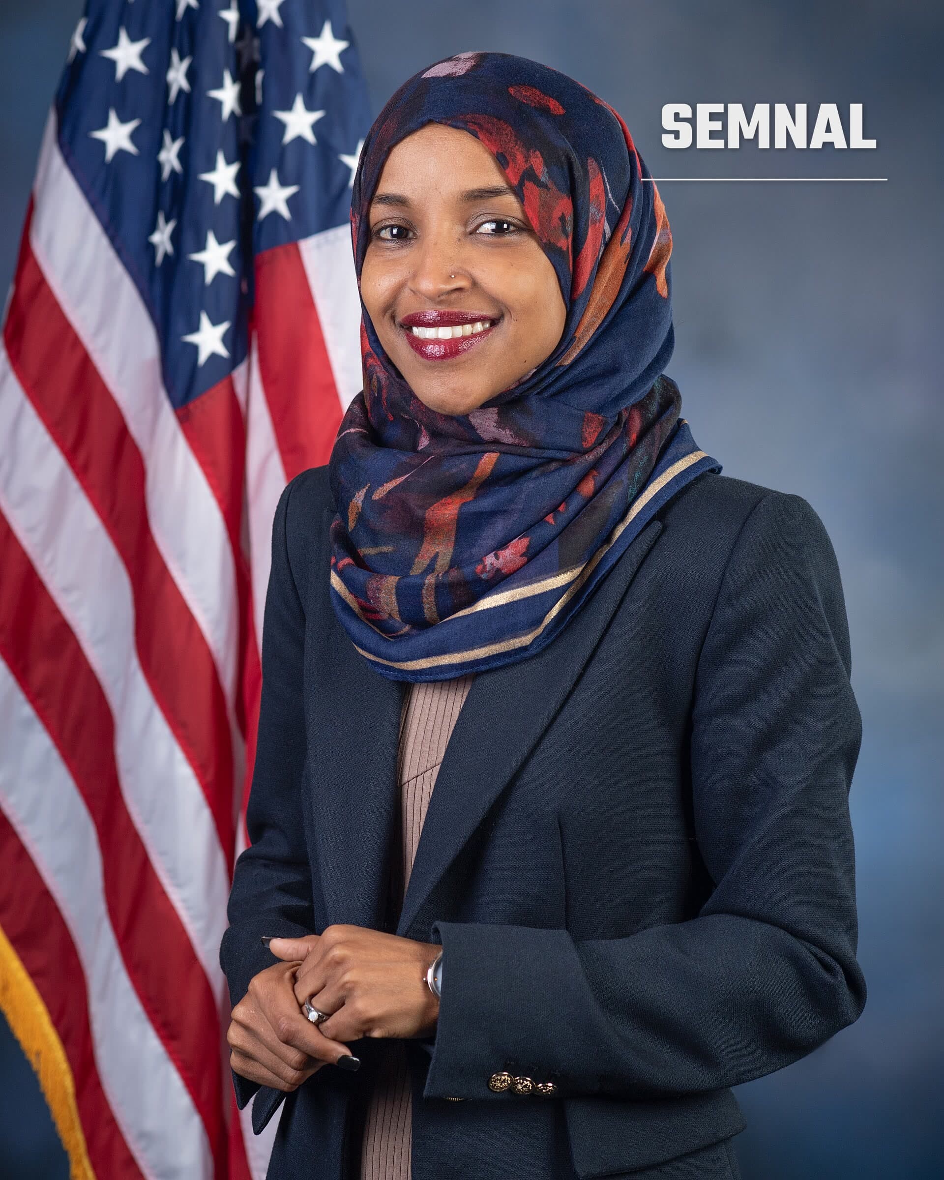 Ilhan Omar zâmbește într-un portret în fața unui steag al Statelor Unite, purtând o eșarfă colorată și un sacou bleumarin. (Foto: Kristie Boyd; U.S. House Office of Photoraphy)
