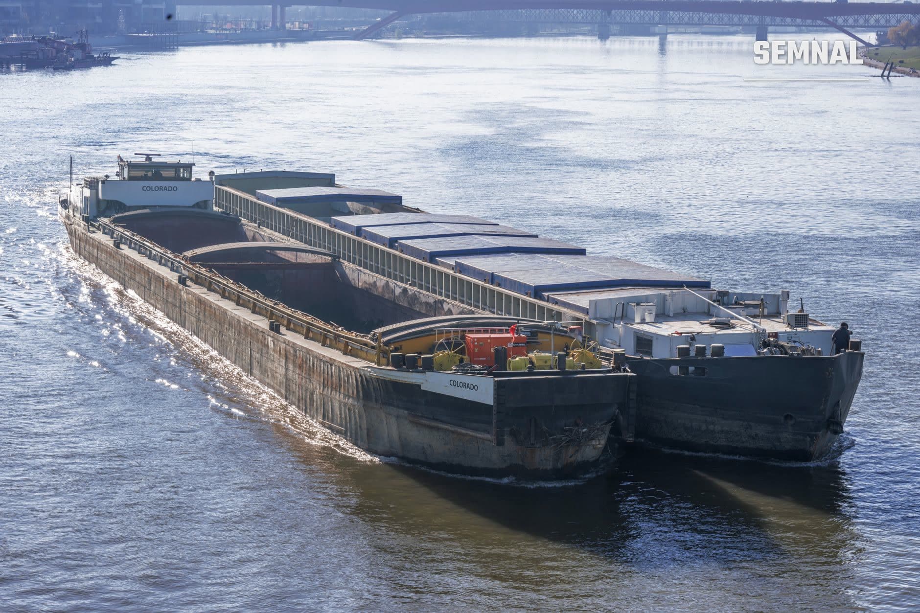 Barcă de transport fluvial Colorado pe Dunăre, încărcată cu cărămizi în compartimente deschise, tracționată de o navă-împingător, cu poduri și infrastructură portuară în fundal. (Foto: Miguel Cuenca / Pexels)